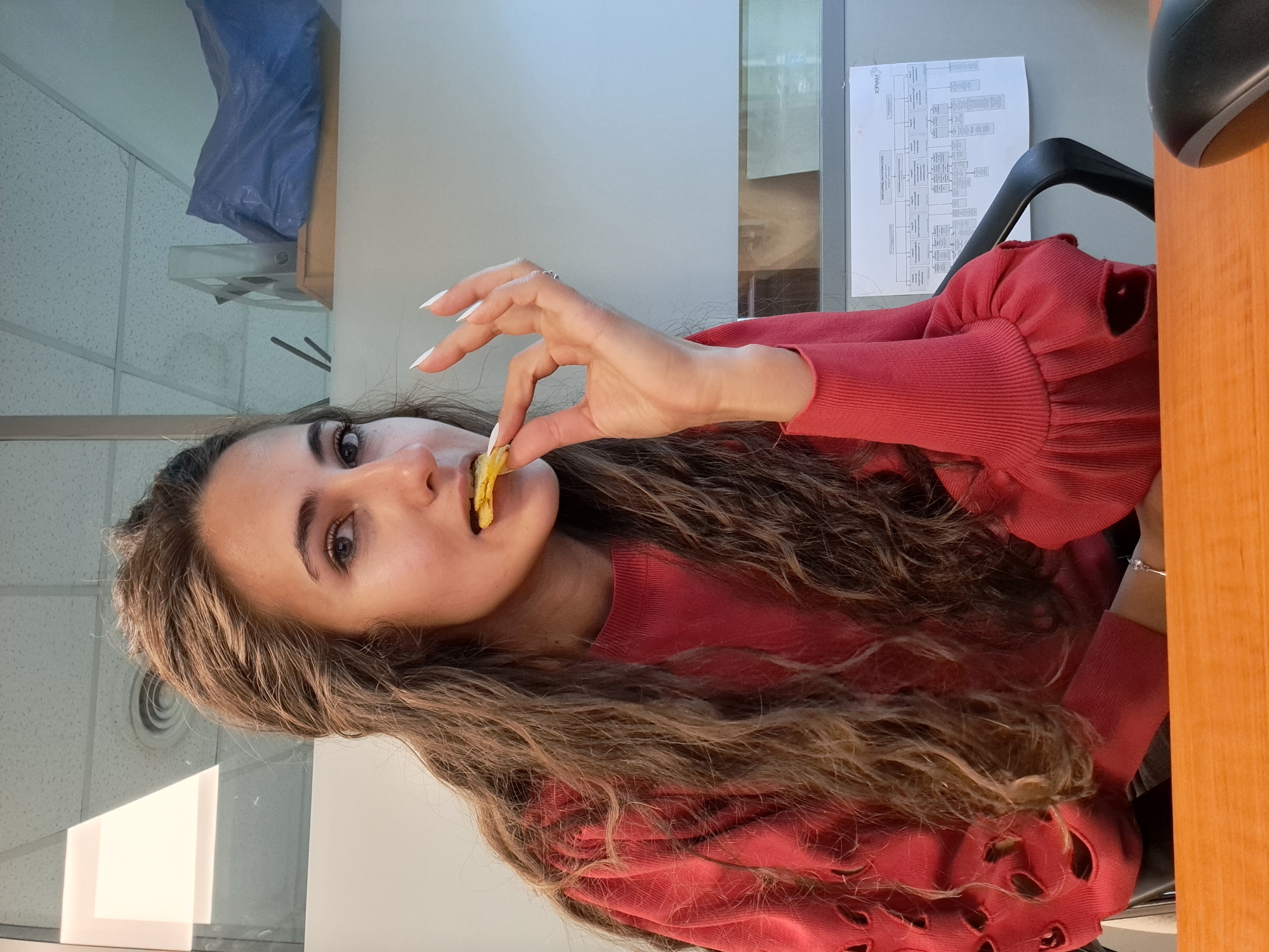 The image features a young woman with long, wavy light brown hair, wearing a red long-sleeved top, looking towards the camera. The image is vertically oriented, showing her upper body. She is holding a crinkled, yellow food item, resembling a potato or plantain chip, to her mouth with her right hand, suggesting she is about to eat it or is in the process of eating. Her hand has long, white manicured nails and a silver ring on her ring finger. A silver bracelet is visible on her left wrist.
The setting appears to be an office or cubicle space. In the background, to the left, a white grid ceiling is visible, along with a blue bag hanging from an overhead fixture. To the right, a wooden desk surface, a black office chair, and a document taped to a wall or partition can be seen. The document contains text and diagrams, possibly an organizational chart. Visible text fragments from the document include "WHISK" (partially) and "XHAM" (mirrored, possibly indicating the original text is reversed). Part of a computer mouse is also visible on the desk. The lighting is consistent with an indoor, artificially lit environment. Her expression is neutral, and the activity seems to be a casual snack break. The provided context states the location is Sofia, Bulgaria.