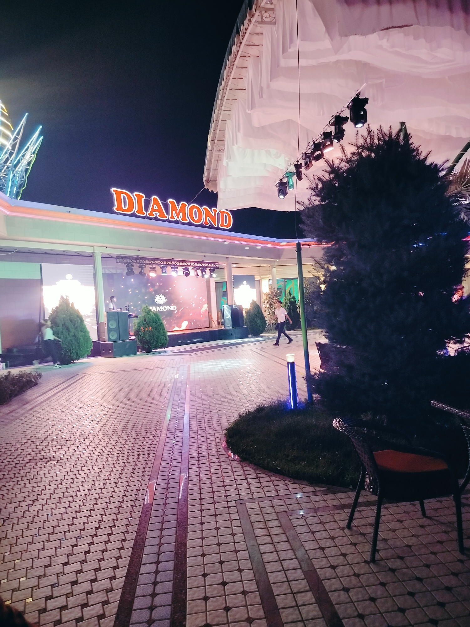 This image depicts an outdoor scene at night, likely at a venue or entertainment establishment in Aksukent, Kazakhstan. The most prominent feature is a large, illuminated sign spelling "DIAMOND" in orange neon letters, situated above an open-air stage. The stage features a large LED screen displaying a graphic with the "DIAMOND" logo, and a person, possibly a DJ, is visible behind a setup of speakers. Several spotlights are suspended above the stage, illuminating the area.
The foreground of the image shows a paved walkway constructed with light-colored square tiles, interspersed with darker, rectangular accent tiles. A dark, evergreen tree dominates the right side of the frame, with its branches obscuring parts of the background. In front of the tree, an outdoor chair with a dark frame and an orange cushion is visible, facing away from the camera.
To the left of the stage, more trees and architectural elements are visible, including a building with a white canopy or awning. The overall atmosphere is festive and brightly lit, characteristic of a night-time public gathering or event. The pavement reflects the colorful lights from the stage and signage, creating a vibrant and dynamic ground-level illumination.