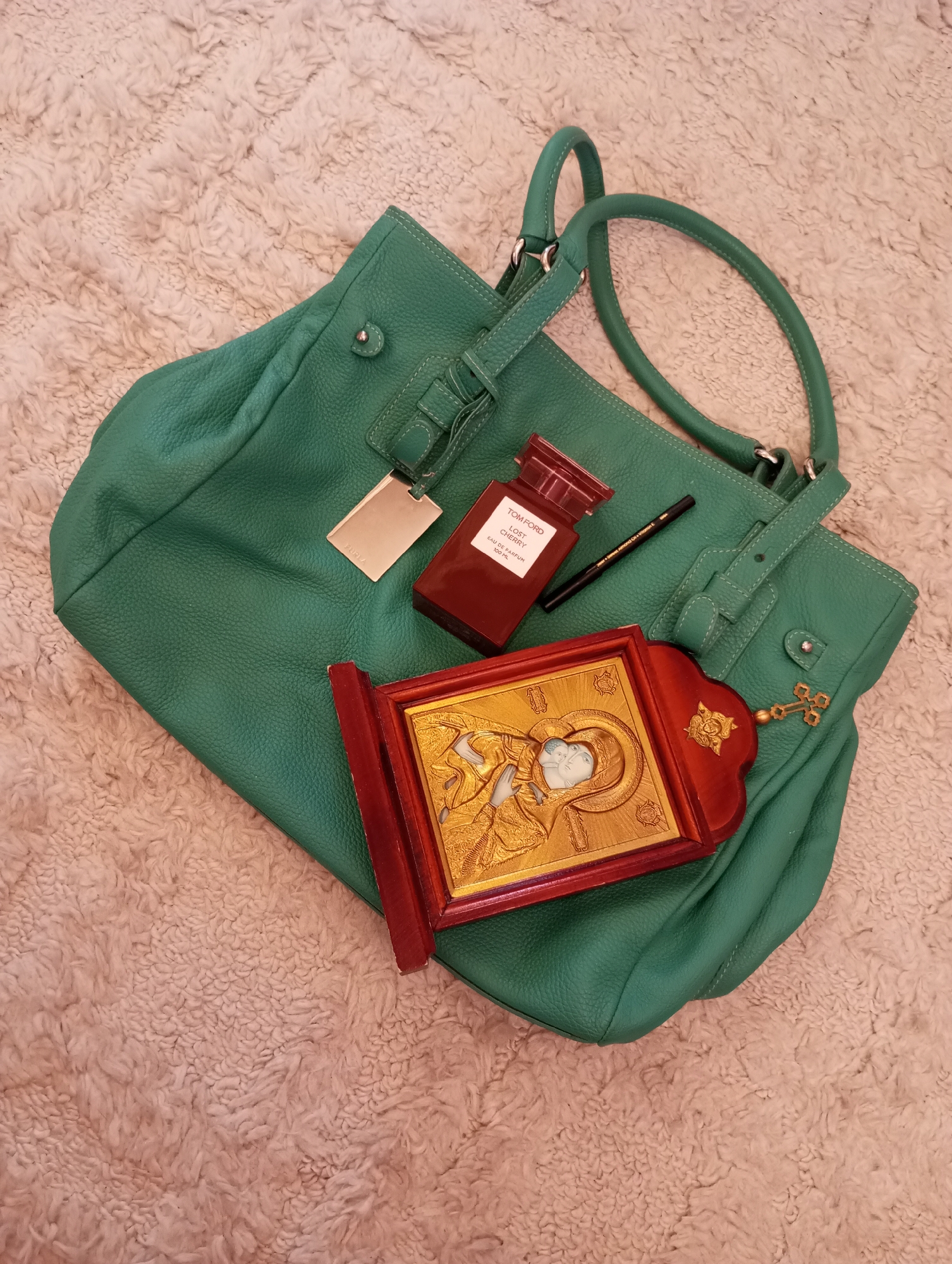 The image shows a green leather tote bag resting on a fuzzy, light-colored surface. On top of the bag, there is a bottle of Tom Ford Lost Cherry perfume, a black eyeliner pencil, and a framed Orthodox icon with a depiction of the Virgin Mary and infant Jesus in gold relief. A small gold cross charm is attached to one of the bag's straps. The location information, Chaikovsky, Russia, does not appear in the image itself.