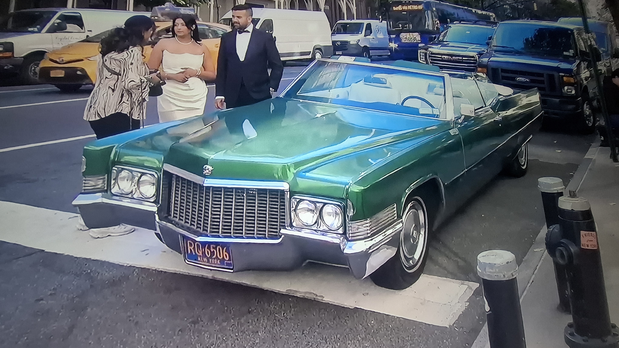 A vibrant green vintage Cadillac convertible occupies the foreground, parked near a sidewalk in an urban environment. The car bears a license plate reading "RQ-6506 NEW YORK." Adjacent to the car, three individuals are positioned on the sidewalk. Two of them, a woman in a strapless white dress and a man in a black tuxedo, stand close to each other, suggesting a formal event or celebration. The third individual, standing slightly apart, is engaged in conversation with the woman in the white dress.
Behind the Cadillac, a variety of vehicles are visible, including white vans, a yellow taxi cab, and a blue bus. These vehicles further emphasize the urban setting. The streetscape is flanked by sidewalks and road markings, indicating a standard city street layout. To the side of the Cadillac, a black fire hydrant and bollards are anchored to the sidewalk. The environment is illuminated by daylight, suggesting an outdoor scene during daytime hours.