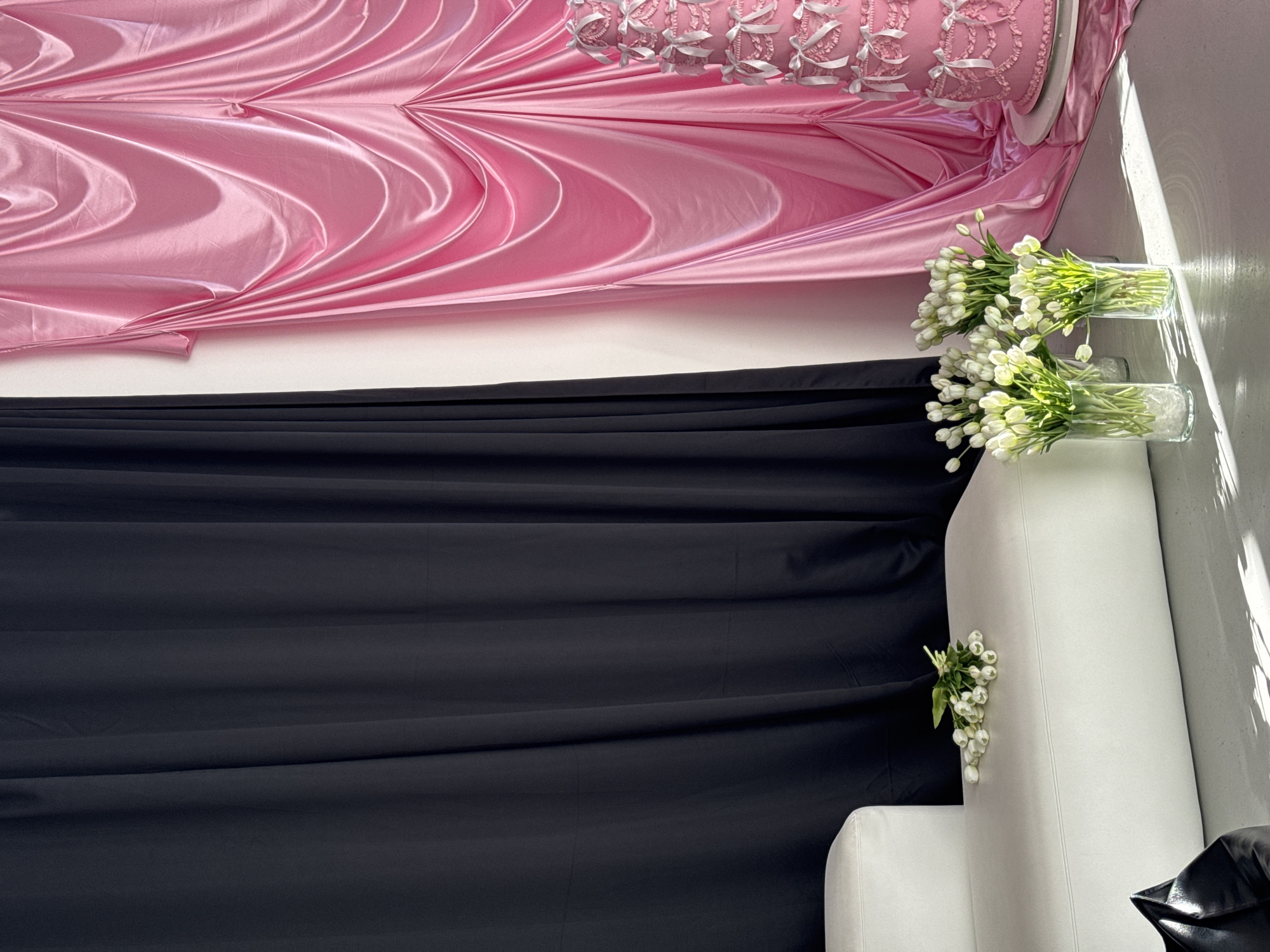 A brightly lit indoor composition features draped fabrics in shades of pink and black against a white wall. The pink fabric, appearing satin-like, cascades from the top of the frame, adorned with decorative trim along the edge. Below it, a swath of black fabric hangs, occupying the middle ground.
To the right, against the white wall, sit two clear glass vases containing bunches of white tulips. A white upholstered chair or sofa is positioned below the flowers, with a smaller bouquet of white flowers placed on its side. Sunlight streams in from the right, casting a shadow on the white wall and the furniture. A shiny black object is visible at the bottom right corner of the frame. The location is in the city of Unknown, Unknown.