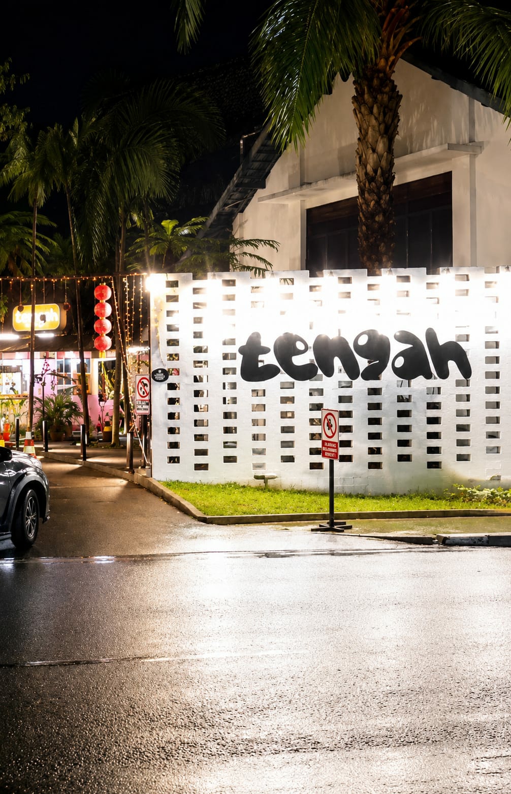 A night-time street-level, medium shot captures a wet asphalt road reflecting the lights of a venue in Medan, Indonesia. A dark-colored vehicle is partially visible on the left. Beyond the road, a white brick wall with rectangular openings is illuminated, featuring the black, bubbly text "tengah" in large lettering. Two red and white circular signs with a red diagonal bar are posted on black stands in front of the wall. To the left of the wall, a row of glowing red lanterns hangs above a building with various businesses and decorative string lights. Palm trees with textured trunks are visible against the dark sky, some with long fronds casting shadows. The ground is covered in grass and a paved curb separates it from the road. The overall scene suggests a vibrant, possibly entertainment-focused, area in the city at night.