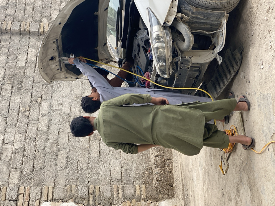 The image depicts an outdoor automotive repair scene in Kandahar, Afghanistan. Three individuals are present around a white sedan with its hood open and front bumper removed, revealing the engine compartment and chassis. A young male, dressed in a light blue/grey shalwar kameez, stands with arms raised, actively working on the engine using a manifold gauge set with connected blue and yellow hoses. Another young male, wearing an olive green shalwar kameez and dark sandals, stands to the left of the working individual, observing the repair. A third person's arm and torso, clad in a brown shirt, are partially visible behind the male in light blue, suggesting involvement in the repair. On the ground near the feet of the person in green, a coiled yellow cable and two light-colored plastic or fiberglass panels are visible. The background features a weathered brick wall, partially covered with a rough cement render. The ground is a mix of concrete and dirt.