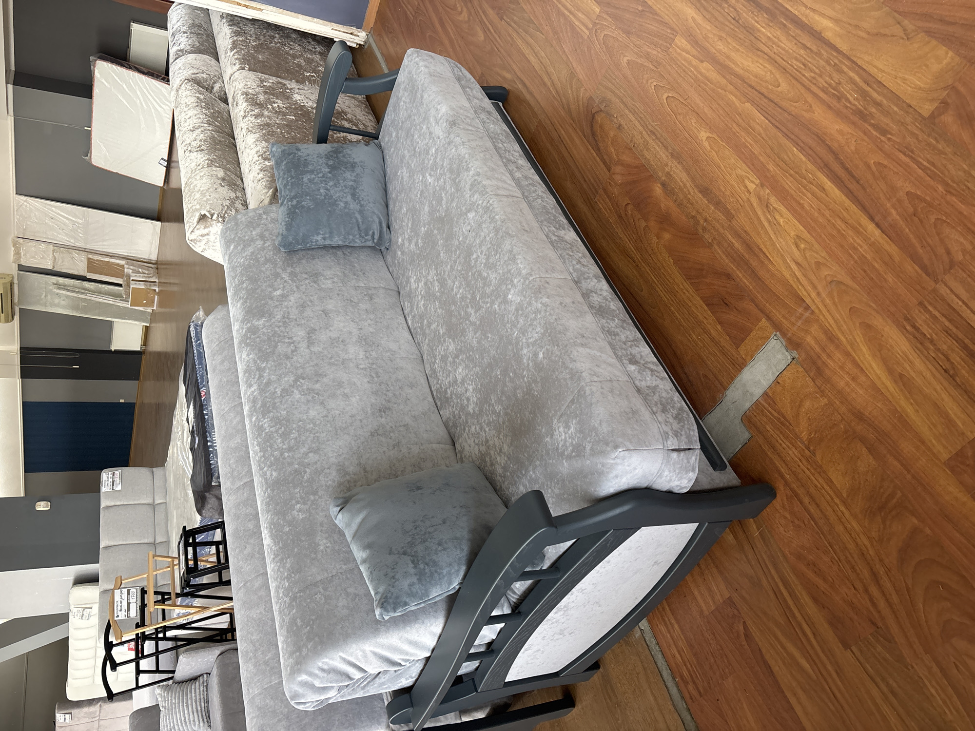 In a furniture store in Zaporizhzhia, Ukraine, a light grey sofa-bed with a textured fabric is displayed on a polished wooden floor. The sofa has dark grey wooden armrests and is adorned with two light grey cushions. In the background, other furniture items, including mattresses and sofas, are visible, along with elements of the store's interior such as walls, lighting, and air conditioning units.