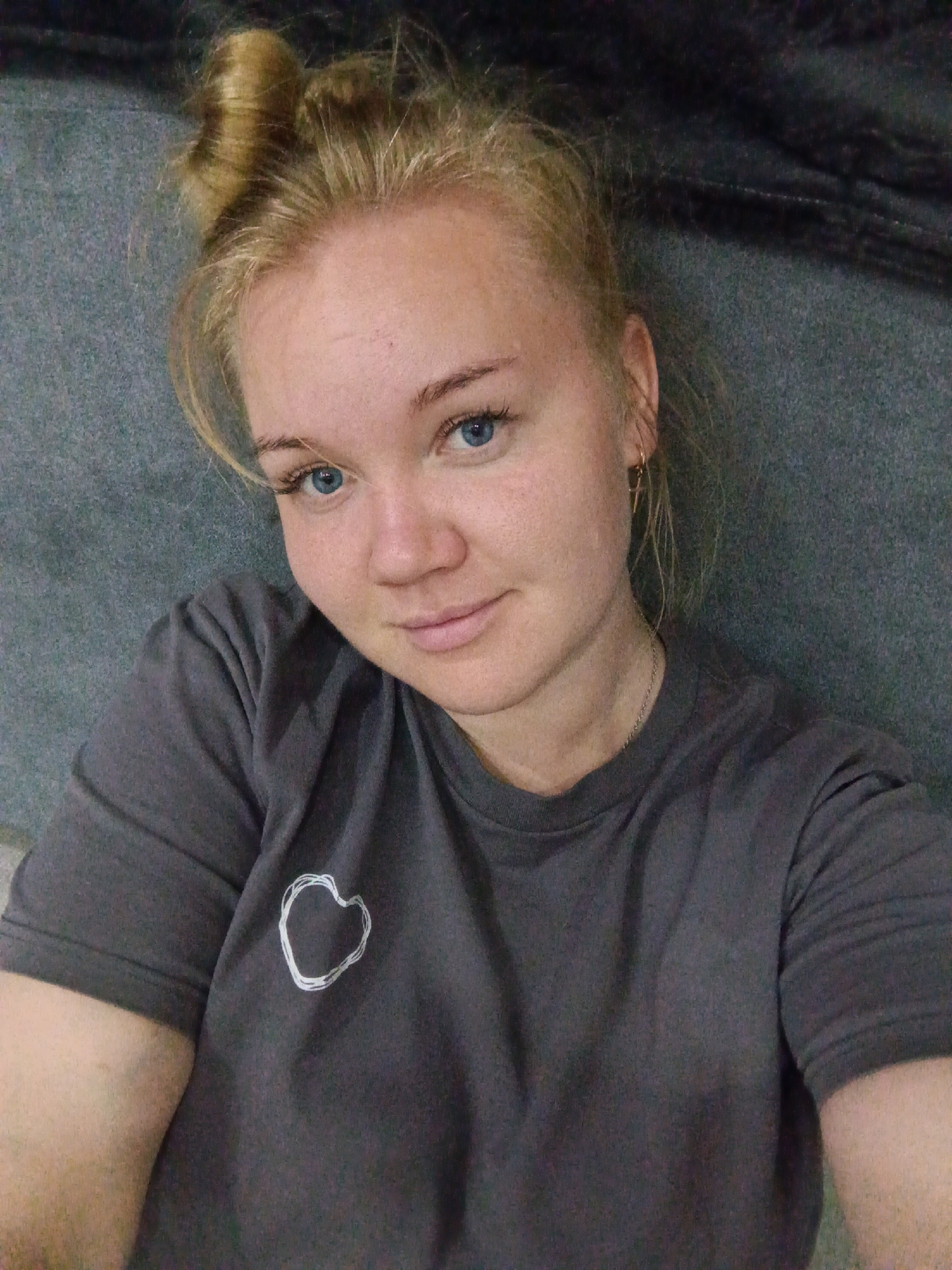 A blonde woman is lying down and looking at the camera. She is wearing a dark gray t-shirt with a white heart outline on it and a silver necklace. Her hair is pulled up into a messy bun. She is also wearing a small hoop earring in her right ear. The background appears to be a gray textured surface, possibly a couch or bed. There is also a dark fabric draped behind her. The lighting is natural and soft, creating a casual and relaxed atmosphere. The woman is located in Foky, Russia.