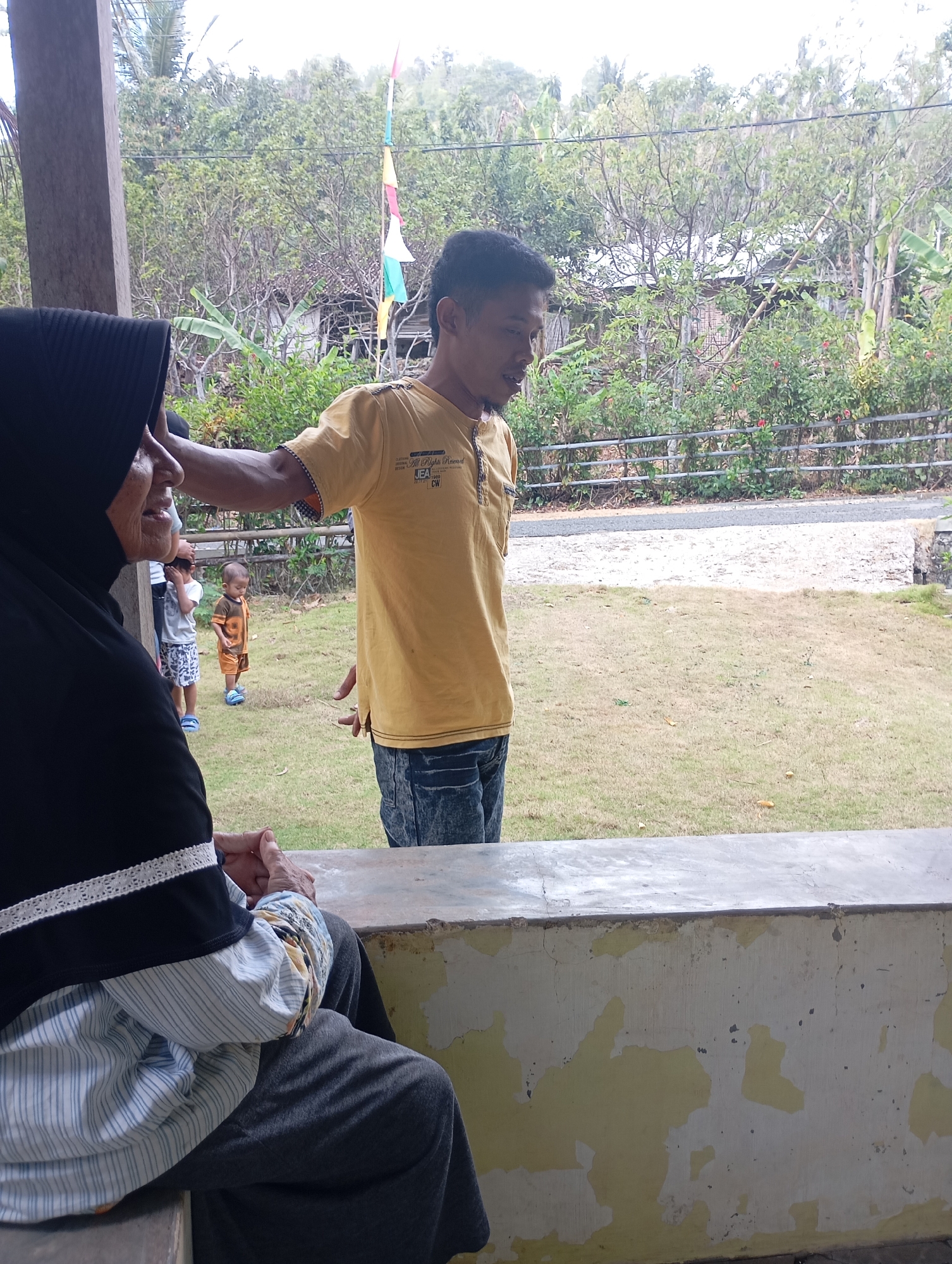 In Ketepung, Indonesia, a man in a yellow t-shirt and blue jeans stands with his right arm extended, seemingly interacting with an older woman wearing a black hijab and a striped shirt, who is seated and facing away from the camera. Behind them, two young children are visible: one in a tan and orange outfit with the number "9" on it, and another wearing a white t-shirt and patterned shorts. The setting appears to be an outdoor area with a grassy lawn and trees in the background. A weathered concrete structure is in the foreground.