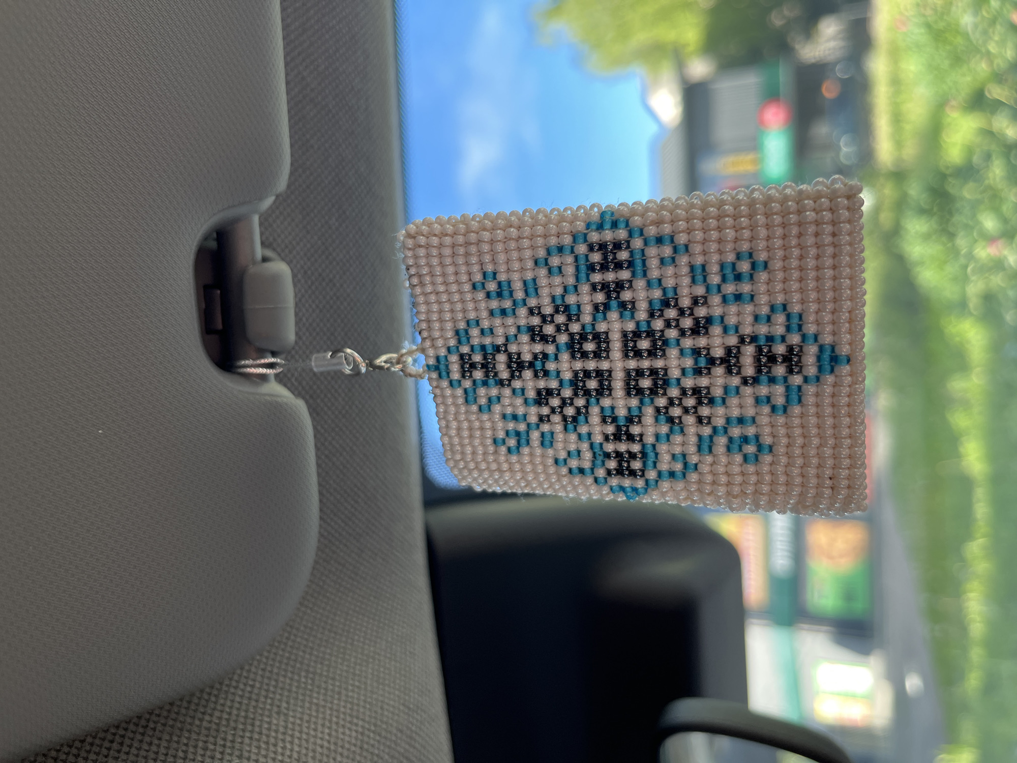 A close-up shot shows a beaded car air freshener hanging from the sun visor inside a vehicle. The air freshener is a rectangular piece with a geometric pattern made of white, teal, and black beads. The background is blurred, showing a view through the car's windshield of a sunny day with a blue sky, some trees, and buildings that appear to be in a commercial area. The location is identified as the city of Zhurivka, Ukraine.