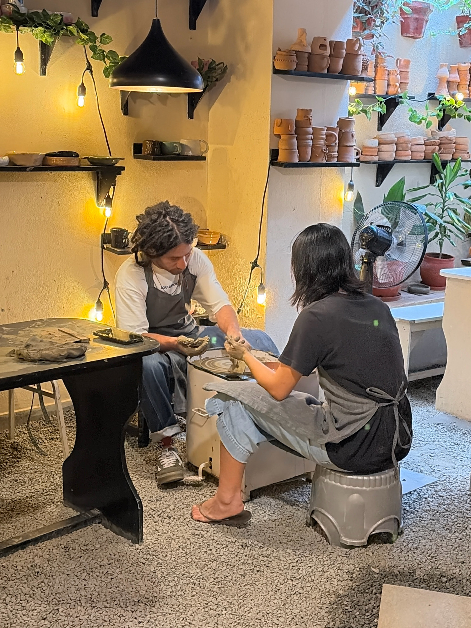The image shows two individuals engaged in pottery work in a studio located in Lalitpur, Nepal. The person on the left, with dreadlocks, is wearing a white shirt and a dark apron over blue jeans, and is seated while working on a pottery wheel. The person on the right, facing away from the camera, has dark hair and is wearing a black t-shirt and a dark apron over blue jeans, and is also working with clay. The environment is a brightly lit workshop with shelves filled with terracotta pottery and plants. Various pottery tools, clay, and finished ceramic pieces are visible throughout the space, including on a table in the foreground and on the shelves. Lighting fixtures are also prominent, with exposed bulbs and a large pendant lamp overhead. A fan is positioned behind the person on the right. The floor is covered with small gravel.