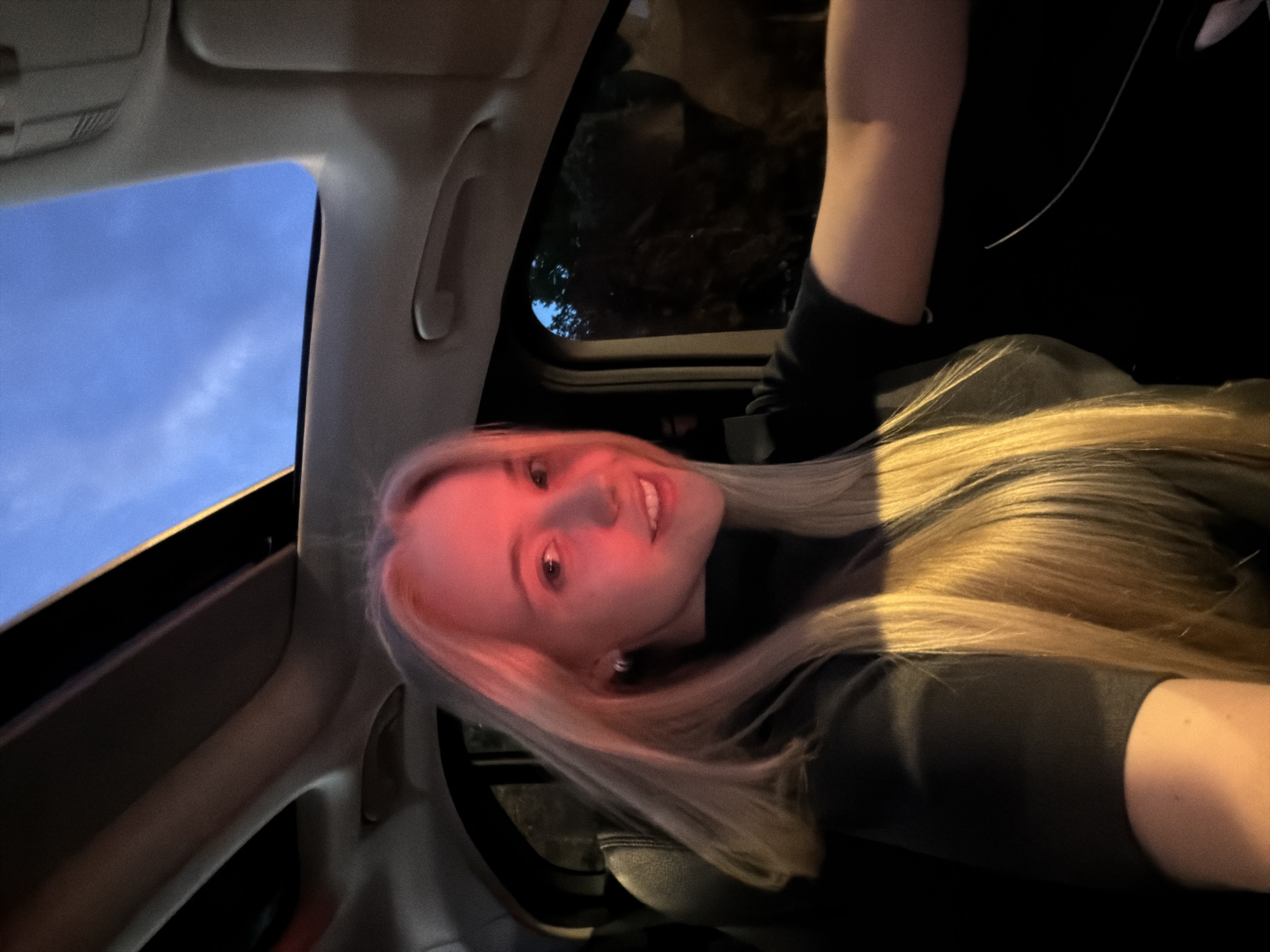 A woman with blond hair is in the passenger seat of a car, likely at night. The car has a sunroof, allowing a view of the sky. The city is unknown.
