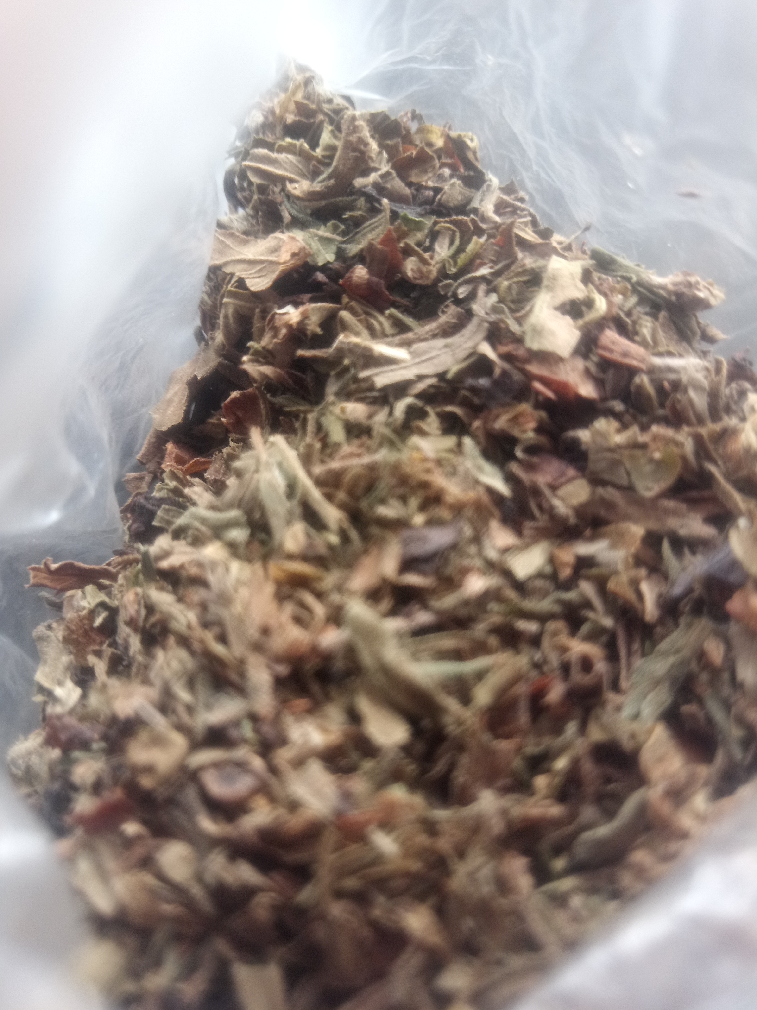 The image displays a close-up view of a pile of dried herbs or spices contained within a translucent plastic bag. The subjects are the dried plant matter, which appears to be a mixture of finely chopped leaves and possibly some stems or flower fragments. The colors range from light brown and tan to muted greens and occasional flecks of darker red or brown. The material is loosely packed, creating a textured surface with visible individual pieces.
The plastic bag is slightly crinkled and shows some diffuse light reflection, indicating it's a typical flexible plastic packaging. The background to the left of the pile is very blurry and appears white or a very light, diffused color, suggesting either a bright light source or a plain, out-of-focus background. The focus is primarily on the dried herbs, with the edges and depth of the pile becoming increasingly blurred towards the top and right of the frame.
There is no discernible activity or event taking place; the image is a still life of the dried material. No people are visible. There are no clear indicators of time of day, weather, or specific location cues, beyond the fact that it is likely indoors due to the lighting and the use of a plastic bag. No text is visible in the image. The overall impression is one of an ingredient or a collected natural product, presented for examination or storage.