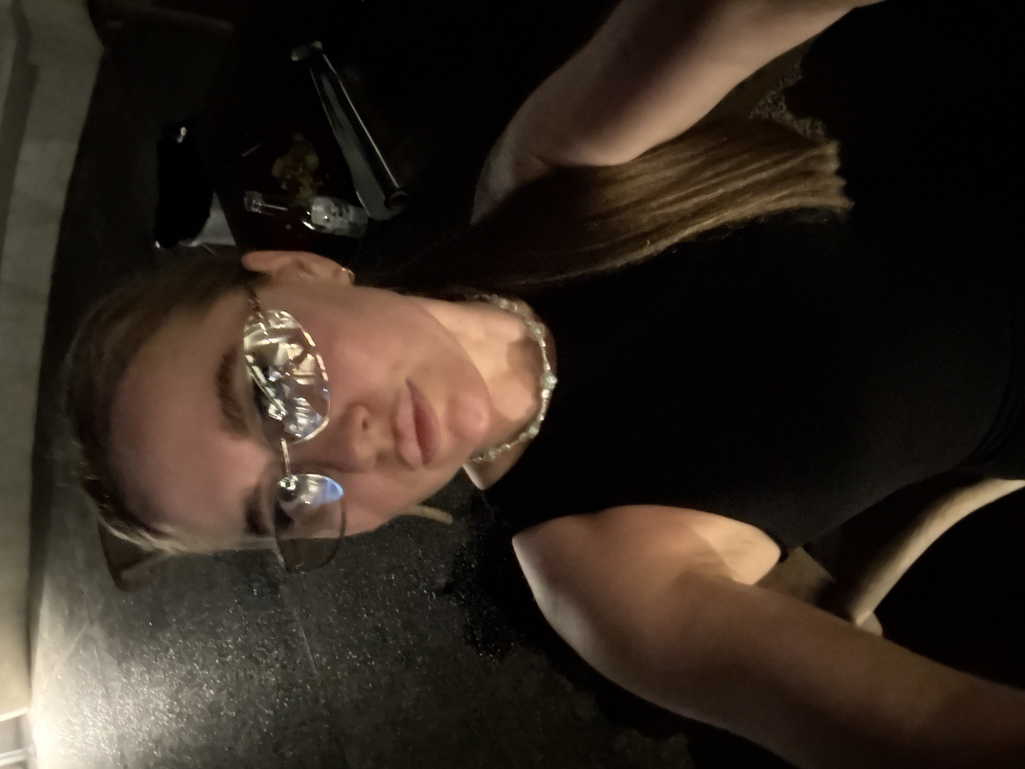 The image shows a woman with blonde hair wearing glasses and a beaded necklace. She is wearing a black top. The setting appears to be a dim or dark location, possibly indoors. There are some objects visible in the background, including what looks like silverware. No information about Jinhua, China can be extracted from this image.