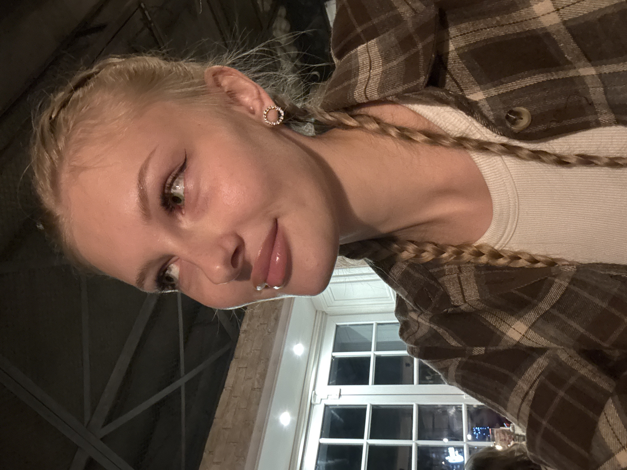 A young woman with blonde hair styled in braids, wearing a plaid shirt and a white t-shirt, is captured in a medium shot. She has a lip piercing and a circular earring with small stones. She is looking away from the camera to her left, with a slight smile. The background includes large windows with white frames, illuminated by lights, and a glimpse of brickwork. The ceiling structure is visible in the upper left corner. The lighting suggests an indoor setting, possibly a restaurant or public space, at night. The location is identified as Perm, Russia.