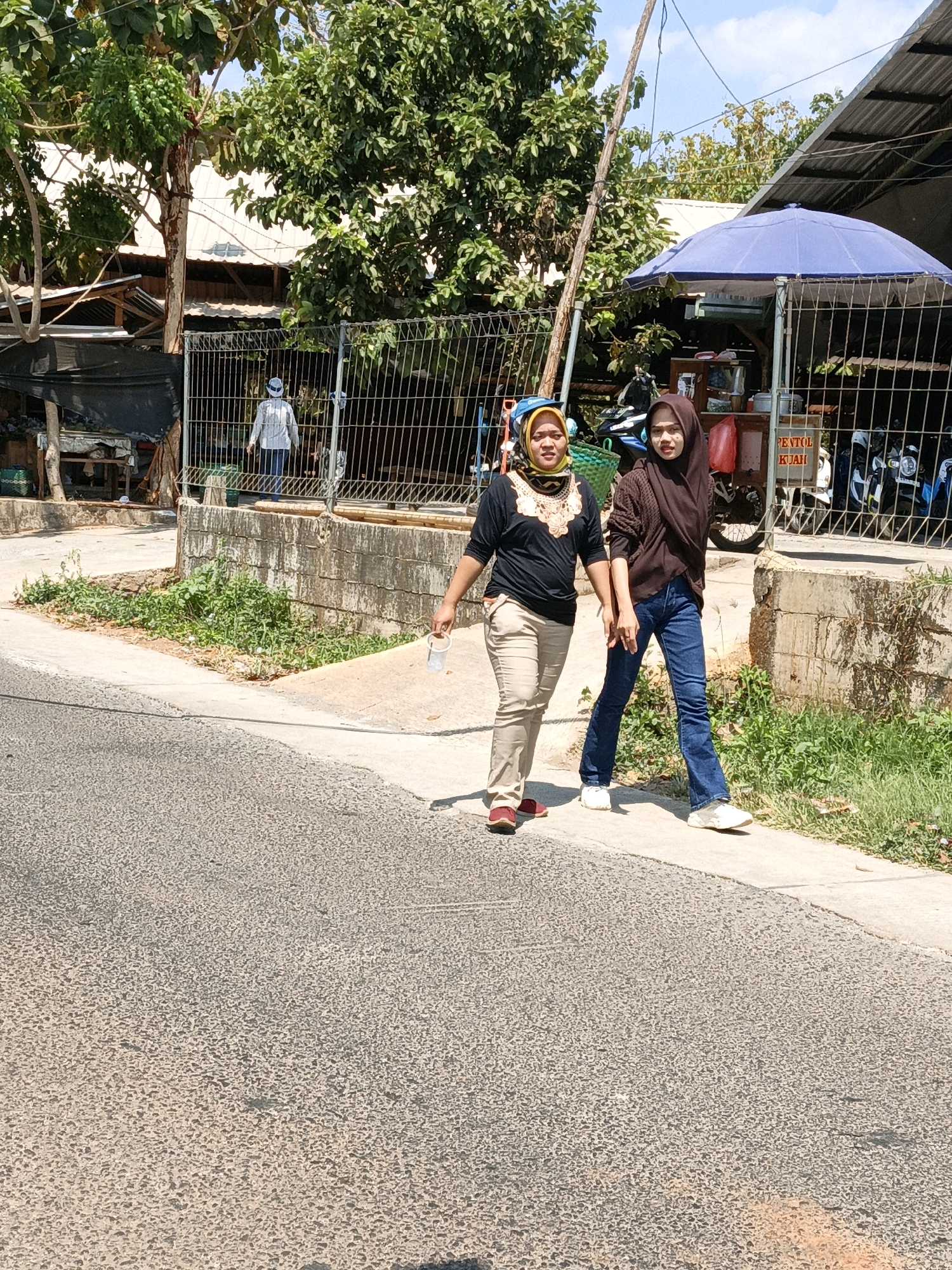 Two women are walking on a paved road next to a concrete sidewalk in Kuwasen, Indonesia. The woman on the left is wearing a black shirt with a patterned neckline, beige pants, and red shoes. She is carrying a clear plastic cup in her right hand. The woman on the right is wearing a brown hijab, a brown long-sleeved shirt, and blue jeans. She is wearing white sneakers. Behind them, there is a metal fence with a stand selling food. Several motorcycles are parked behind the fence. Trees and buildings are visible in the background under a clear blue sky.