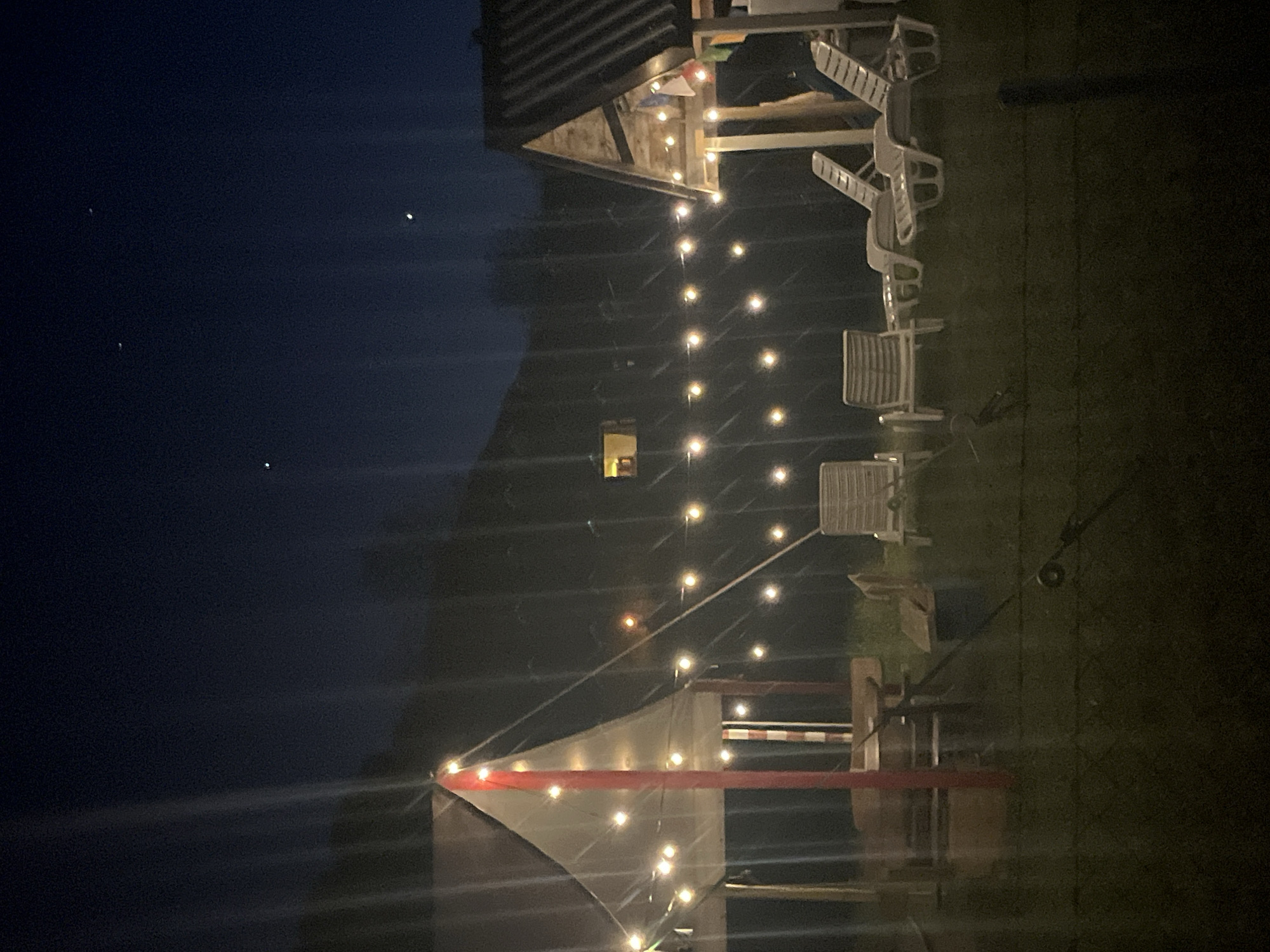 The image depicts a dark outdoor scene, likely at night, with a partial view of a building illuminated by string lights. The building has a visible window and appears to be constructed with wood or similar materials. To the right of the building, there is a grassy area with stacked white plastic chairs. A shade sail, possibly triangular, is visible in the foreground, also adorned with string lights. The background shows a dark sky. The location in Polja, Montenegro, is not directly evident from the image itself. No people are visible.