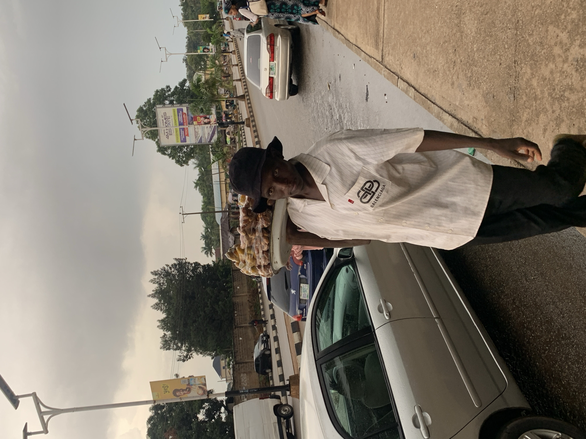A young Black male is captured mid-stride on a cloudy day in Jos, Nigeria, carrying a large platter of food on his head. He wears a black bucket hat and a white striped short-sleeved shirt with a prominent Balenciaga logo on the left breast pocket. His dark pants are visible. The background depicts a street scene with several cars, including a silver sedan in the foreground on the left and a silver sedan further down the street on the right. A blue hatchback is also visible in the middle ground. Buildings and trees line the street, and streetlights are present. The sky is overcast, suggesting potential rain, and the road appears wet. An advertisement banner with the text "YES!" is visible on a lamppost to the left. Another large billboard advertising "4M HOMES ESTATE" is situated further down the street. The overall environment suggests a busy urban setting in Jos.