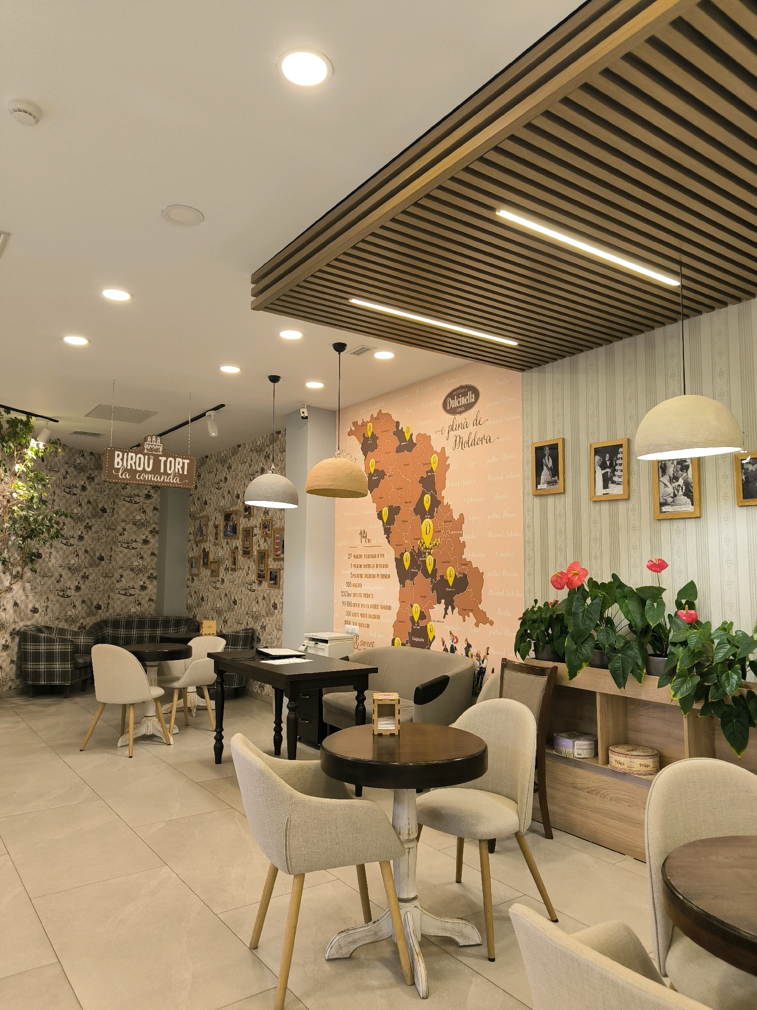 This image depicts the interior of a cafe in Chișinău, Moldova. The cafe features a warm and inviting ambiance with a prominent map of Moldova decorating one wall. The map has various points of interest marked, and text in Romanian, including the cafe's name, "Dulcinella," and phrases like "plina de Moldova" (full of Moldova) and "BIROU TORT la comanda" (cake office to order).
The furniture consists of several round wooden tables, some with ornate bases, surrounded by light-colored upholstered chairs with wooden legs. A dark wood desk is also visible, along with a printer. In the background, a patterned wallpaper covers some of the walls, and a large stone-like wall is partially visible. Framed photographs are hung on the wall above a comfortable-looking plaid sofa.
Potted plants, including red anthuriums, add a touch of greenery and color to the space, arranged on a wooden shelf unit. The ceiling has a modern design with wooden slats and recessed lighting, as well as pendant lights. The floor is tiled with large, light-colored tiles.
The lighting is soft and ambient, contributing to the cozy atmosphere of the cafe. No people are visible in the image, focusing the viewer's attention on the decor and layout of the establishment. The overall impression is of a well-designed and comfortable cafe.