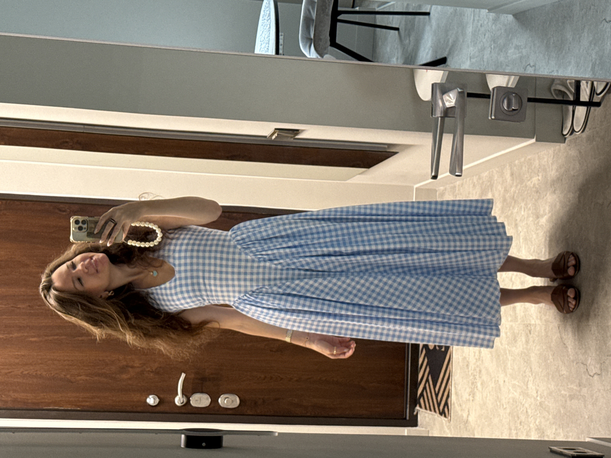 A woman stands in a hallway, taking a mirror selfie. She is wearing a blue and white gingham dress and brown sandals. She has long, brown hair and is smiling at her reflection. The hallway has a wooden door, a tiled floor, and a large mirror. In the reflection, a round table with a chair is visible.