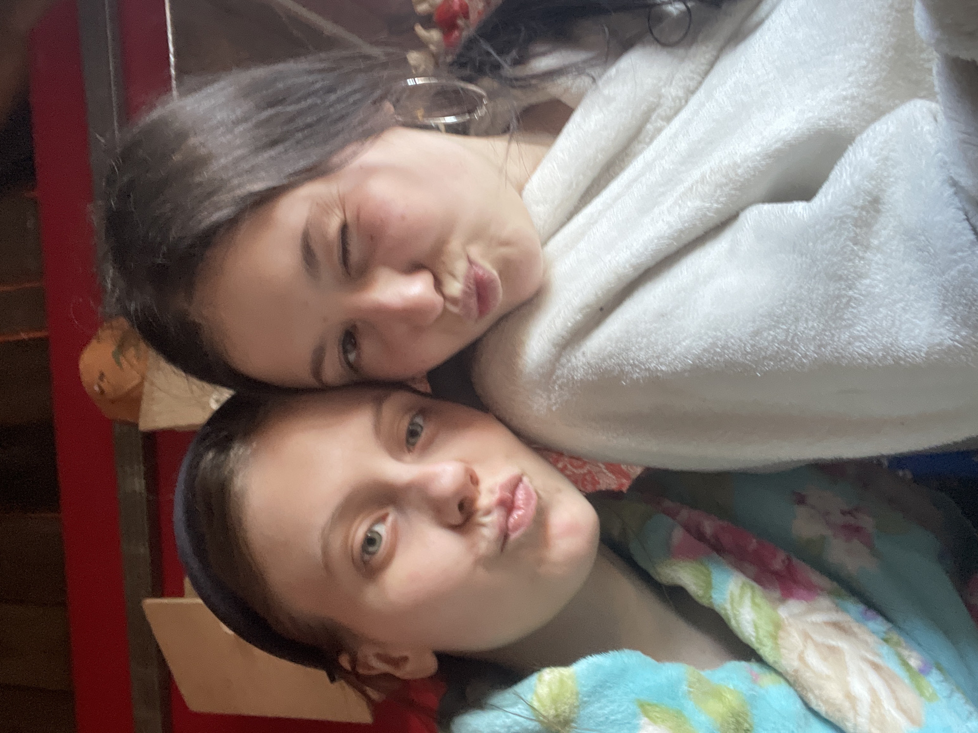 Two young women are posing closely together. The woman on top winks, has dark hair, and wears hoop earrings; she is wrapped in a white blanket. The woman below has light hair and is covered in a patterned blue blanket. Both women are making kissing faces. The location is unclear, and there is no indication it is in Фоки, Russia.