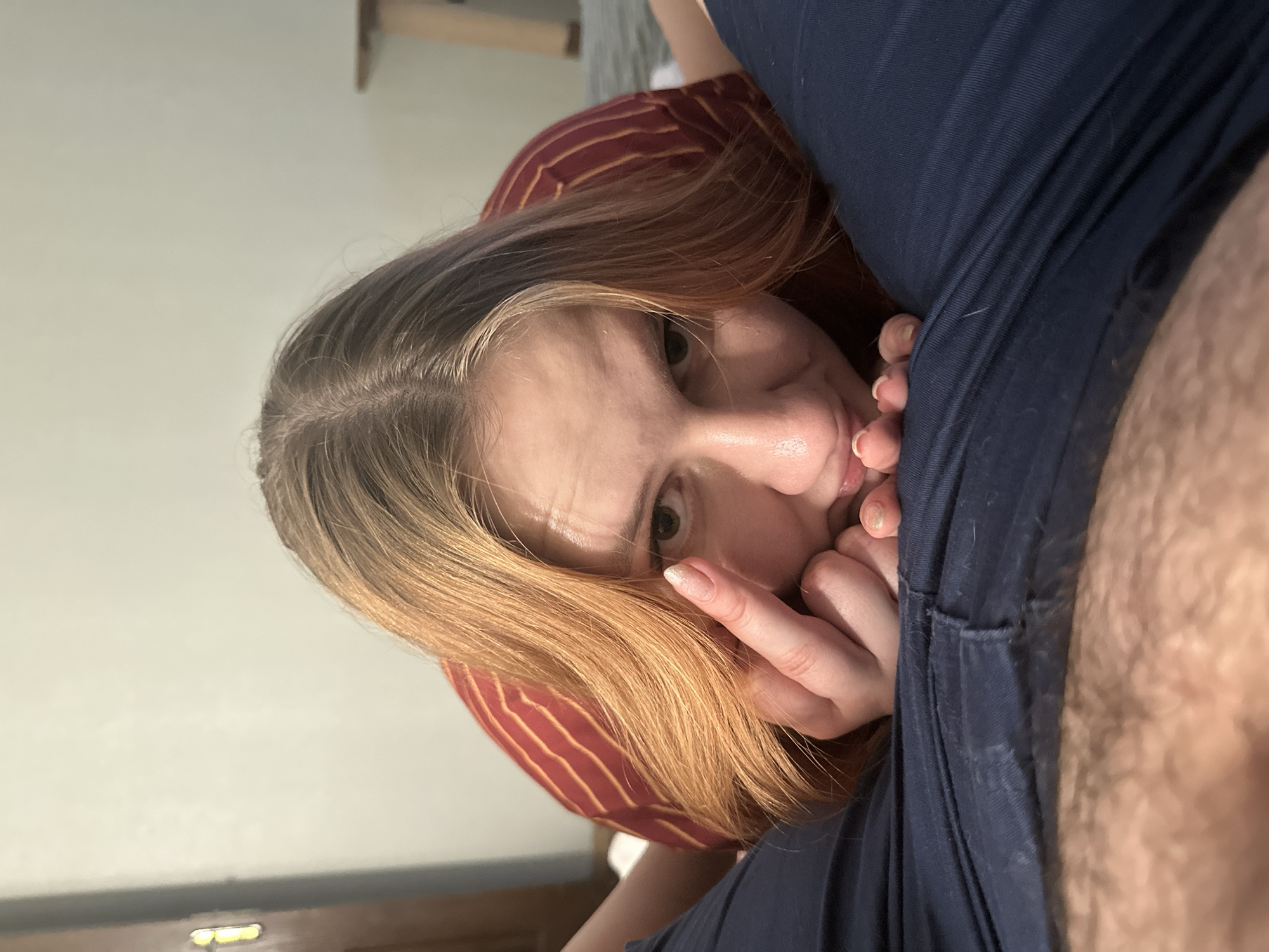 The media file is a close-up, low-angle photograph of a young woman's face. She has blonde hair with reddish undertones and is wearing a red and yellow striped shirt. Her eyes are wide and looking directly at the camera, with a slightly surprised or intense expression. Her hands are near her mouth, with one finger touching her lips. The lighting is soft and appears to be indoor, possibly from a natural light source given the subtle shadows. The background is blurred, but appears to be a domestic setting with a hint of a wall and furniture. The overall mood is intimate and possibly suggestive due to the subject's pose and facial expression. There is no visible text in the image.