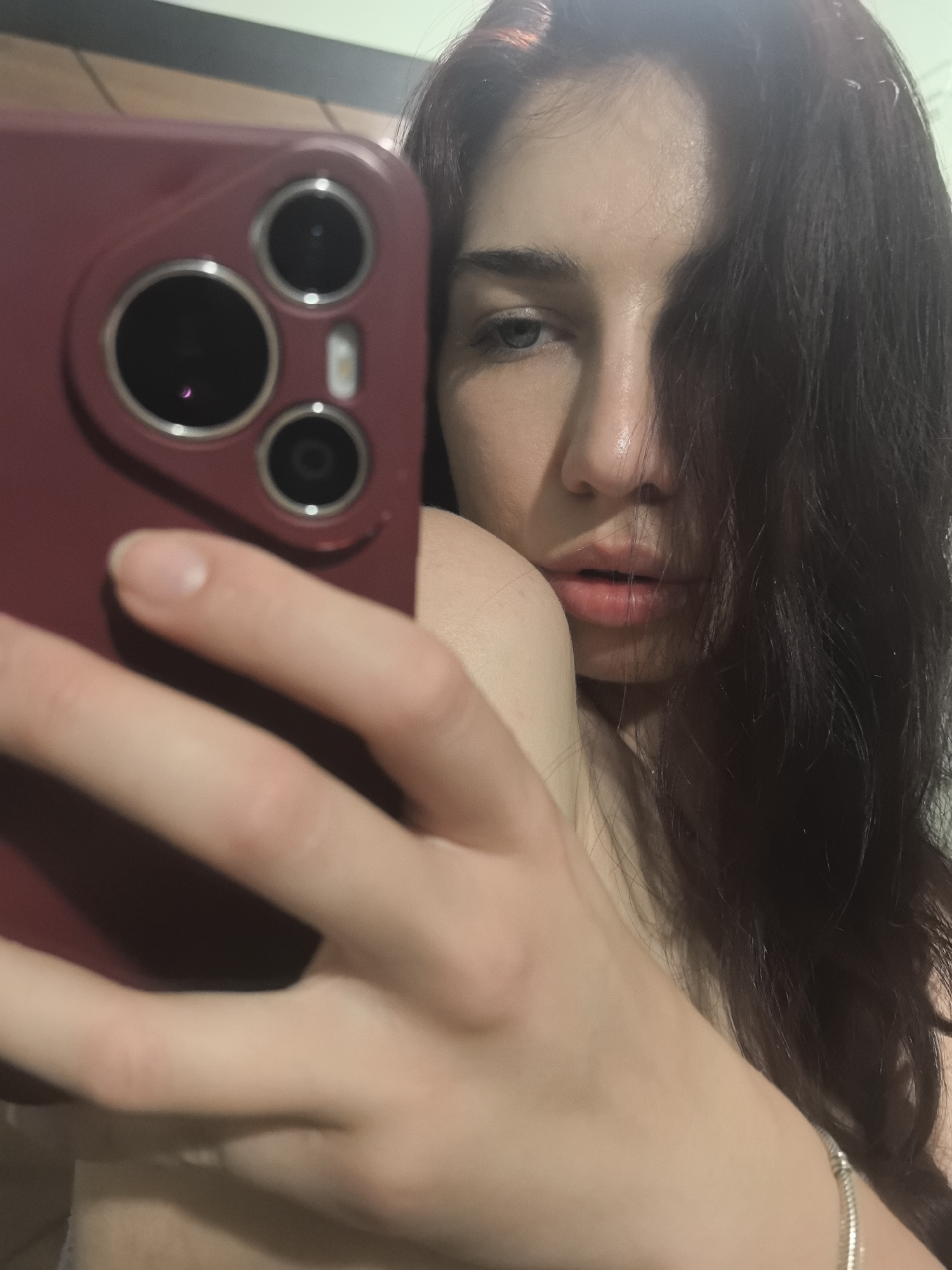 The image is a selfie taken by a woman. She is holding a red phone with three visible camera lenses. The woman has dark hair that covers part of her face. She has fair skin and light eyes. She is wearing a silver bracelet on her wrist. The background is indistinct. The location and city are unknown.