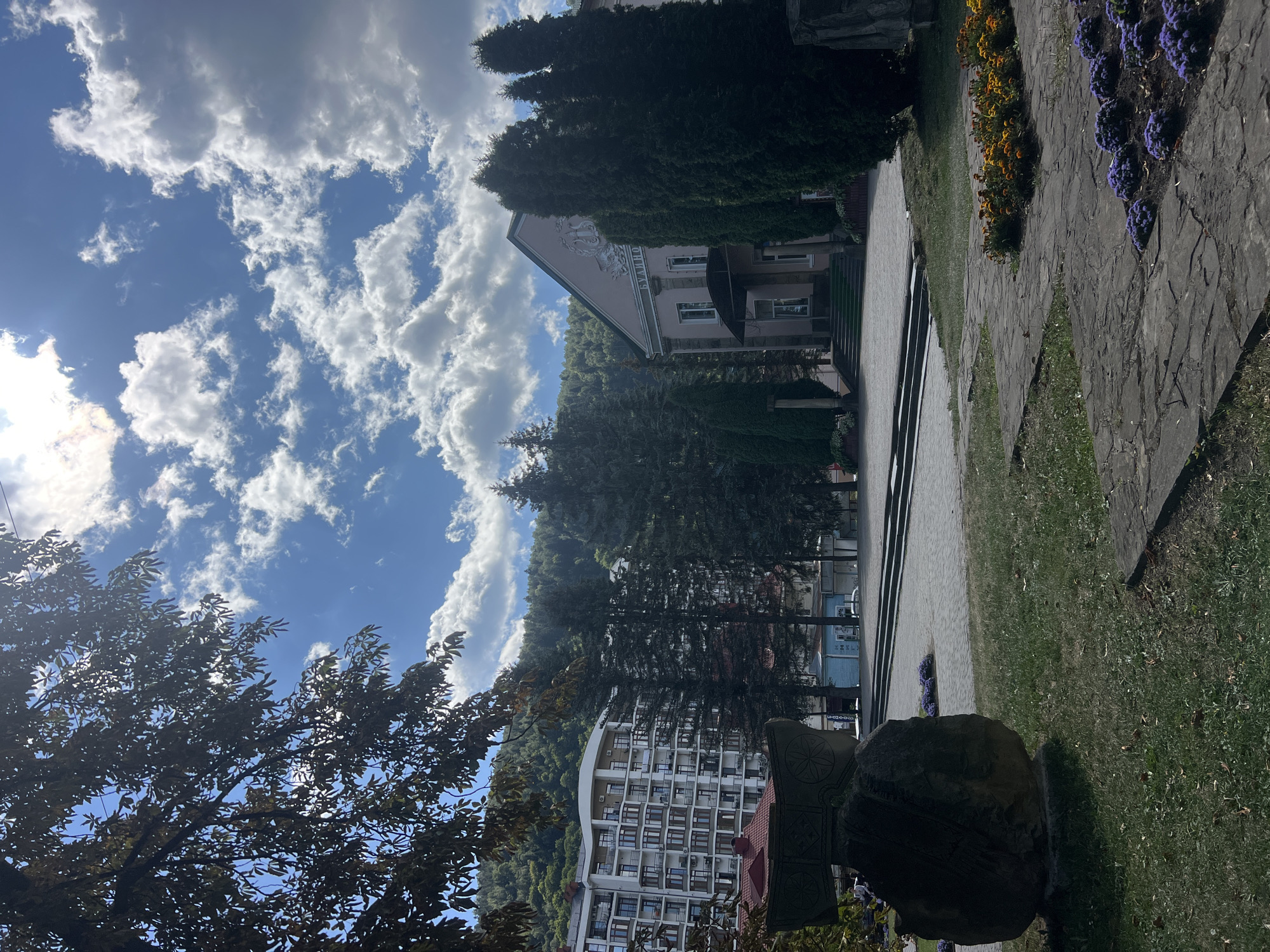 The image depicts a sunlit plaza in Yaremche, Ukraine. In the foreground, a large, weathered stone sculpture sits on a grassy area, with paved pathways leading to a central plaza. Flowerbeds with orange and purple blooms are visible along the pathways.
The plaza is paved with stone and features a tiered set of stairs leading up to a pink building with an arched entrance and decorative elements on its facade. The building has windows and a dark-covered entrance with steps. Tall, dark green coniferous trees are planted in front of the building and to its right.
Behind the plaza, a modern, multi-story white building with numerous balconies is visible, nestled among more tall trees. In the background, a lush green, tree-covered hillside rises under a bright blue sky dotted with white, fluffy clouds. The overall scene suggests a public square in a town surrounded by nature. No people are clearly visible in the image.