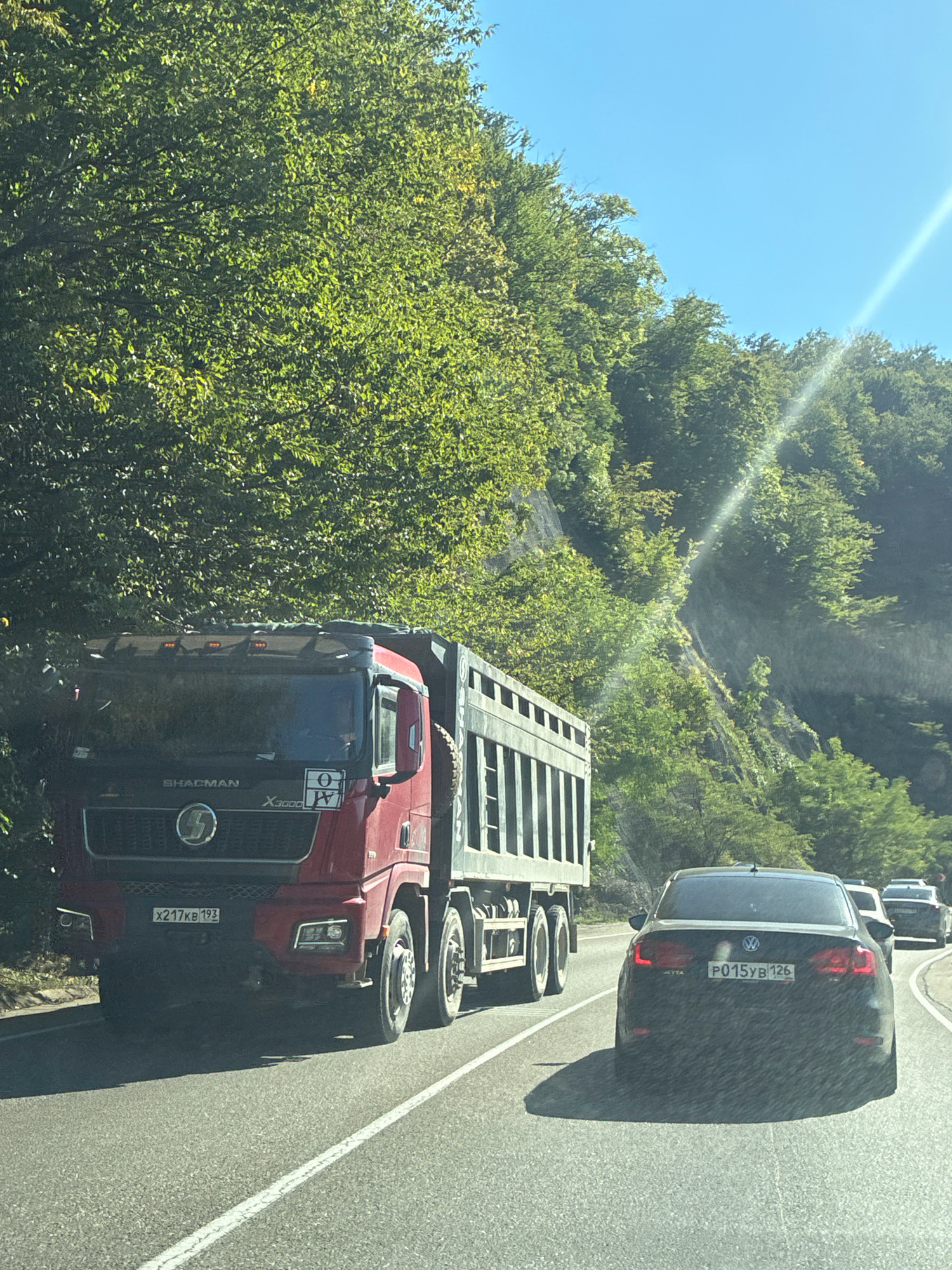A red SHACMAN X3000 dump truck with a license plate ending in 193 is on a paved road, traveling behind a dark Volkswagen Jetta with a license plate ending in 126. The truck is in the left lane, and the Jetta is in the right lane. The road is surrounded by a steep, forested hillside with lush green trees. The sky is bright blue with a sun glare visible on the right side of the image. The scene appears to be captured from a moving vehicle, likely another car on the road. The location is identified as the city of Agoy, Russia.