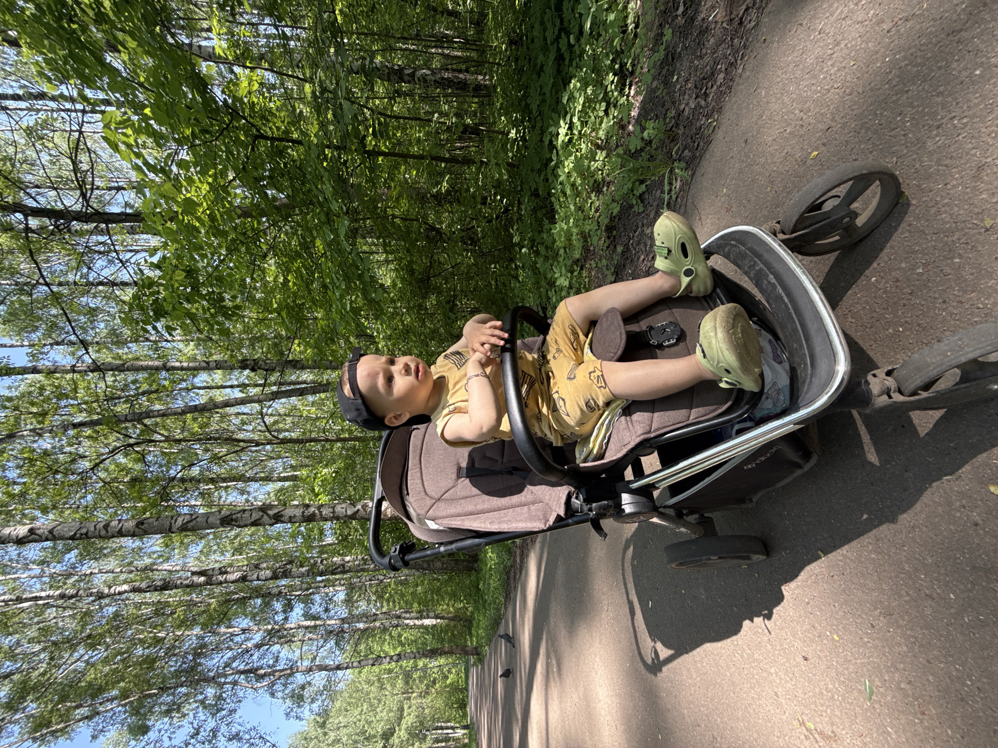 A young child is lying in a stroller on a path. The child is wearing a yellow outfit and light green clogs. The stroller is on a paved path through a park or wooded area. Tall trees line the path. The photo was taken in Заволжье, Russia.