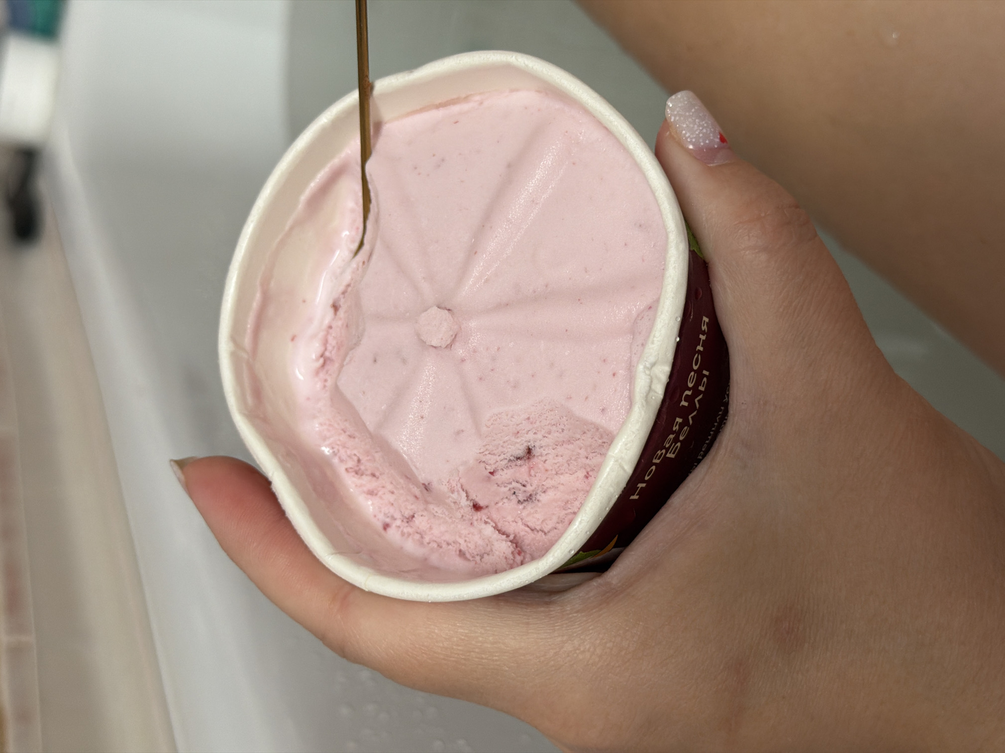 A close-up, top-down shot depicts a person holding a partially eaten container of pink ice cream in their left hand. A spoon is visible, partially inserted into the ice cream. The container appears to be made of paper and has some text in a non-English language (Russian) printed on its side. The person's left thumb and forefinger are visible, with the forefinger sporting a decorative nail design. The background is a blurred, bright white, possibly a sink or counter surface.
Based on the text on the ice cream container, specifically "Новая песня беллы", it can be inferred that the location could be linked to the city of Chaikovsky, Russia, given the Russian text and potential association with the ice cream brand/manufacturer.