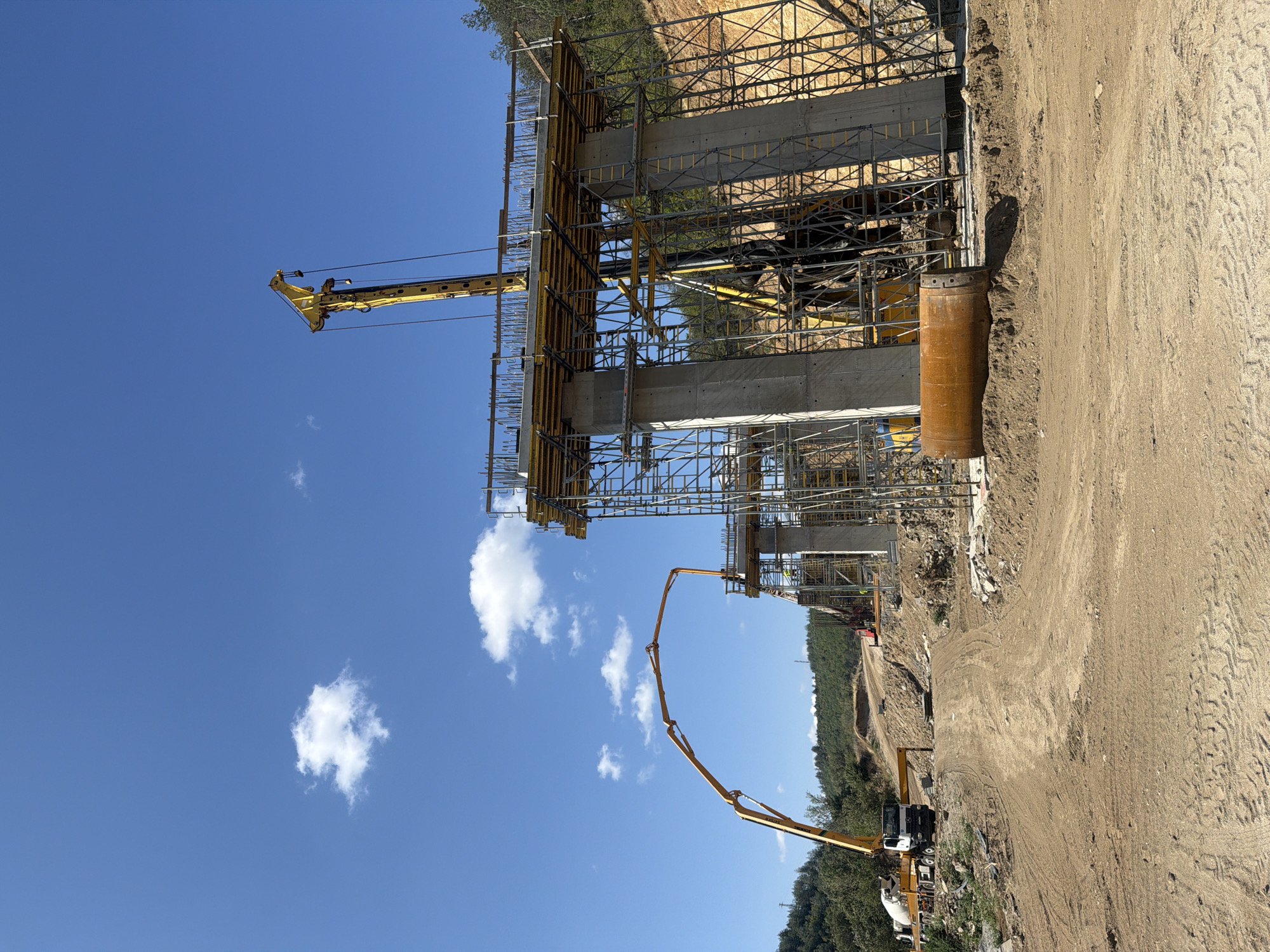 In Kresna, Bulgaria, a large-scale construction project is underway, focusing on the development of a new overpass. The scene is dominated by towering concrete pillars, intricately laced with scaffolding and formwork, indicating the advanced stage of construction. A concrete mixer truck, a common sight at such sites, is positioned on the left, ready to supply the essential material for the project. Adjacent to it, a concrete pump truck extends its articulated arm, poised to deliver concrete to the elevated sections of the overpass. In the background, a dense forest line encircles the construction zone, suggesting a rural or semi-rural setting. The sky above is a clear blue, punctuated by a few fluffy white clouds, implying fair weather conditions conducive to construction work.