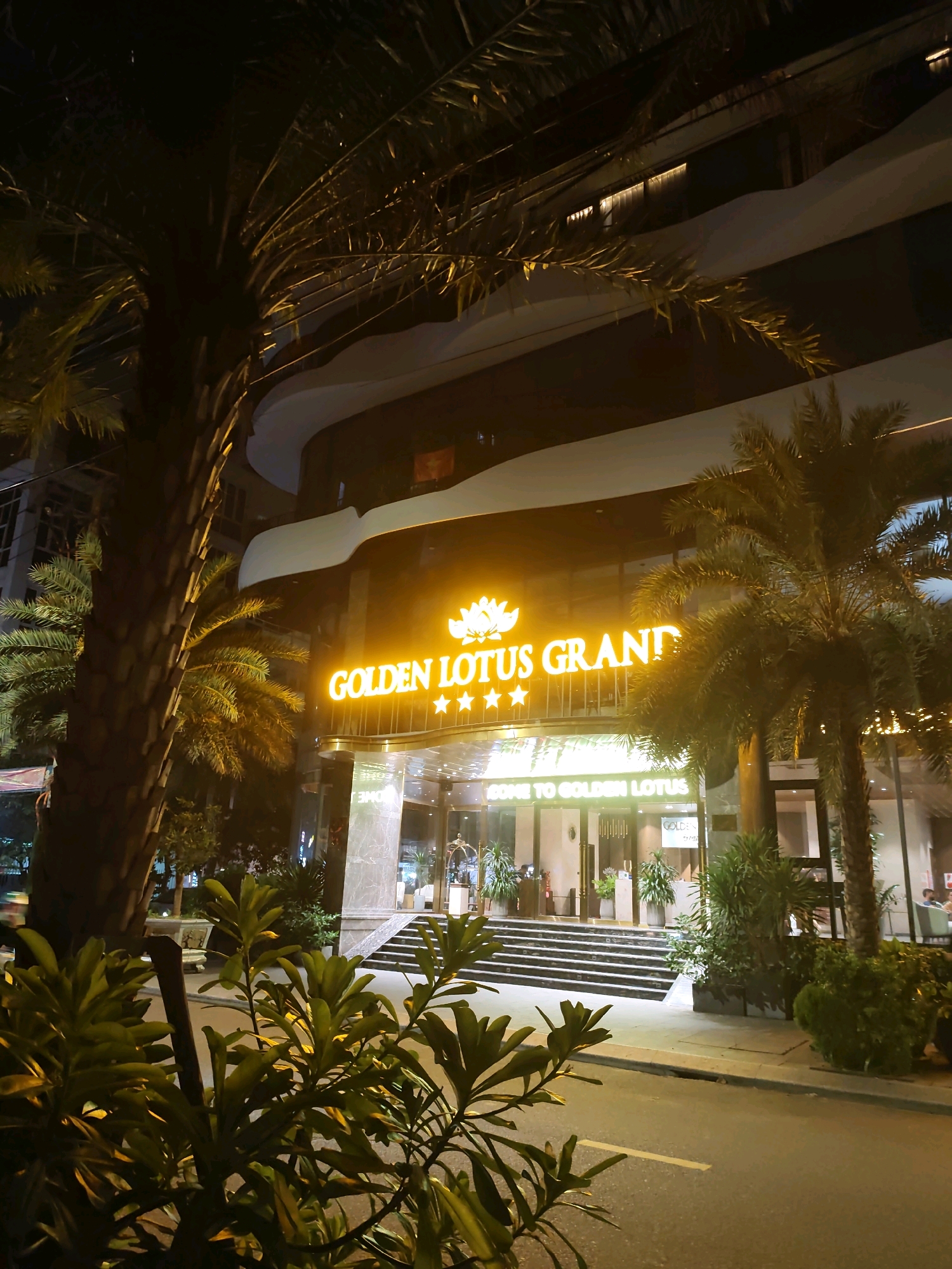 An exterior shot at night shows the entrance to the Golden Lotus Grand hotel. A large, illuminated sign with the hotel's name and a lotus flower logo hangs above the entrance. Palm trees frame the building, and foliage lines the walkway. Stairs lead up to the glass doors of the hotel, and a sign reads "WELCOME TO GOLDEN LOTUS." A street runs in front of the hotel, with a few cars visible in the distance. The scene is lit by the hotel's lights and ambient city light.
