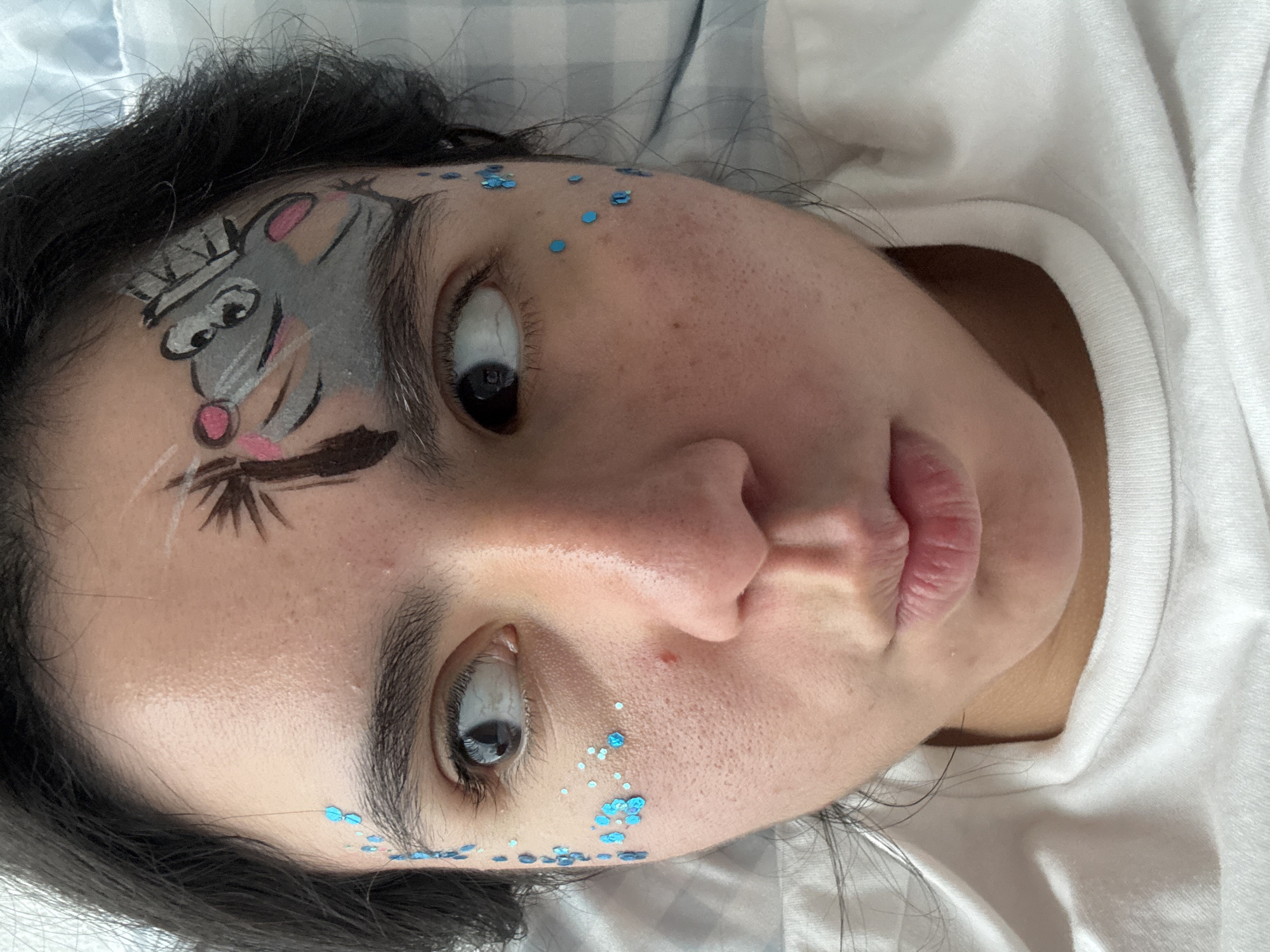 A close-up shot captures a person's face with face paint and glitter. The face paint depicts a cartoonish rodent on the forehead and eyebrows. Blue glitter is applied in clusters near the eyes and temples. The person is wearing a white t-shirt. The background is a light-colored, checkered fabric, possibly a blanket or pillowcase. The image is taken from a high-angle perspective, looking down at the person's face. The lighting is soft and even. The specific location in the city of Austin, United States of America, cannot be determined from the image alone.