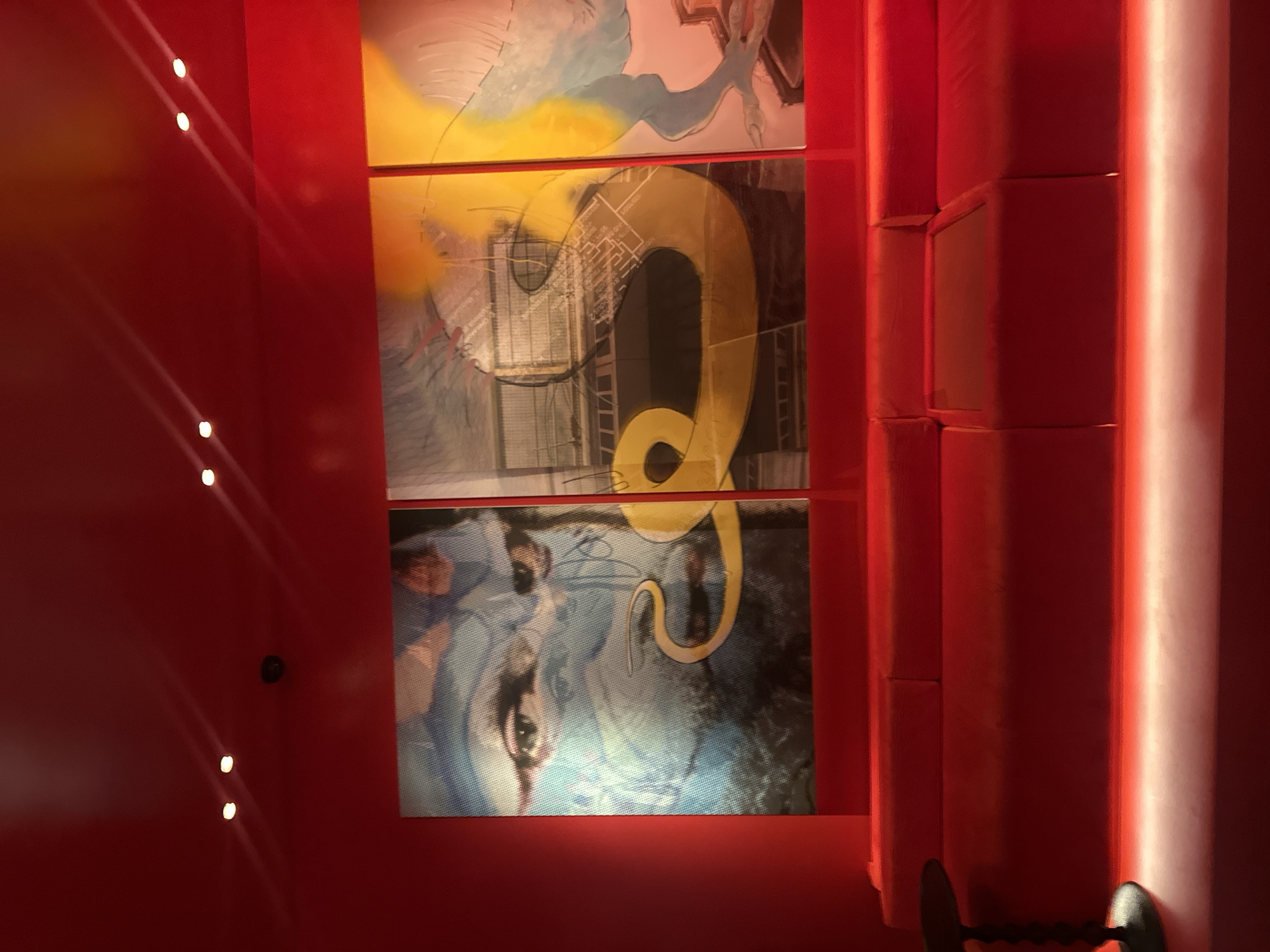 The image shows a photograph of an interior space featuring a multi-panel artwork. The artwork, divided into three horizontal sections, displays abstract imagery with elements that suggest architectural drawings and a human face. The color palette is dominated by reds, yellows, blues, and grays. The artwork is hung on a red wall, and to the right of the art, a structure of stacked, blocky, red forms is visible. A light fixture casts a bright, vertical line along the edge of the structure.
Based on visual features, it is difficult to definitively ascertain the location of the photo in London, United Kingdom.