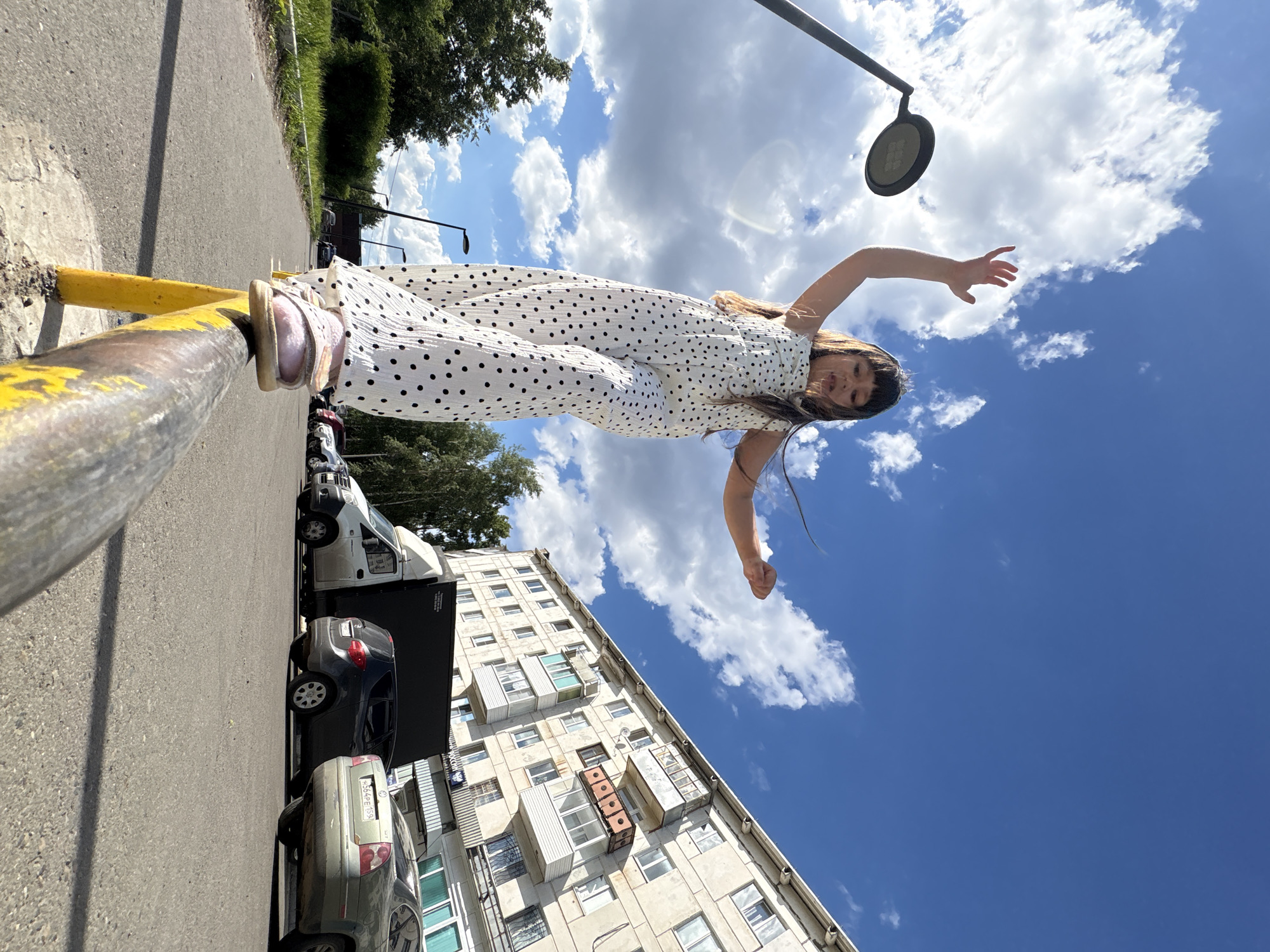 A low-angle, full-body shot shows a young girl walking on a yellow-and-gray metal railing in Chaikovsky, Russia. She is wearing a white jumpsuit with black polka dots and pink sandals. Her arms are outstretched for balance, and her hair is blowing in the wind. The background features a blue sky with scattered white clouds, a streetlamp, trees, a multi-story apartment building, a delivery truck, and several parked cars. The image is taken outdoors on a sunny day.