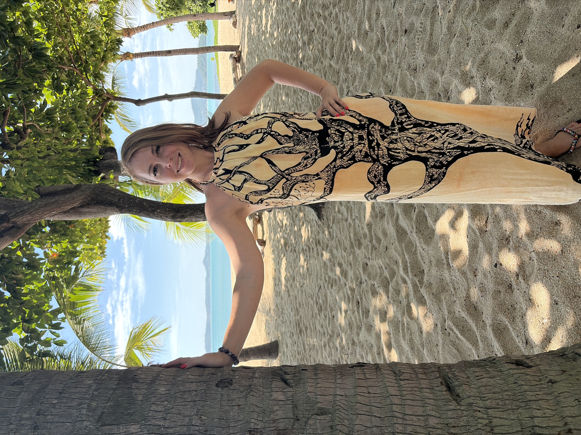 A blonde woman with fair skin and a light tan stands on a sandy beach in Ko Samui District, Thailand. She is wearing a sleeveless, halter-neck dress with a light yellow base and a black tree-like pattern. The dress reaches her ankles. Her right hand rests on her hip, and her left arm is extended and leaning on a palm tree trunk to her left. She wears a dark beaded bracelet on her left wrist and flip-flops on her feet. The woman is smiling at the camera.
The background shows a tropical beach scene with more palm trees and lush green foliage. The sand is light brown and textured with ripples. Beyond the beach, the turquoise ocean meets a clear blue sky with scattered white clouds. In the distance, a hilly island is visible. The lighting suggests it is daytime, with dappled sunlight filtering through the leaves of the trees, casting shadows on the sand.