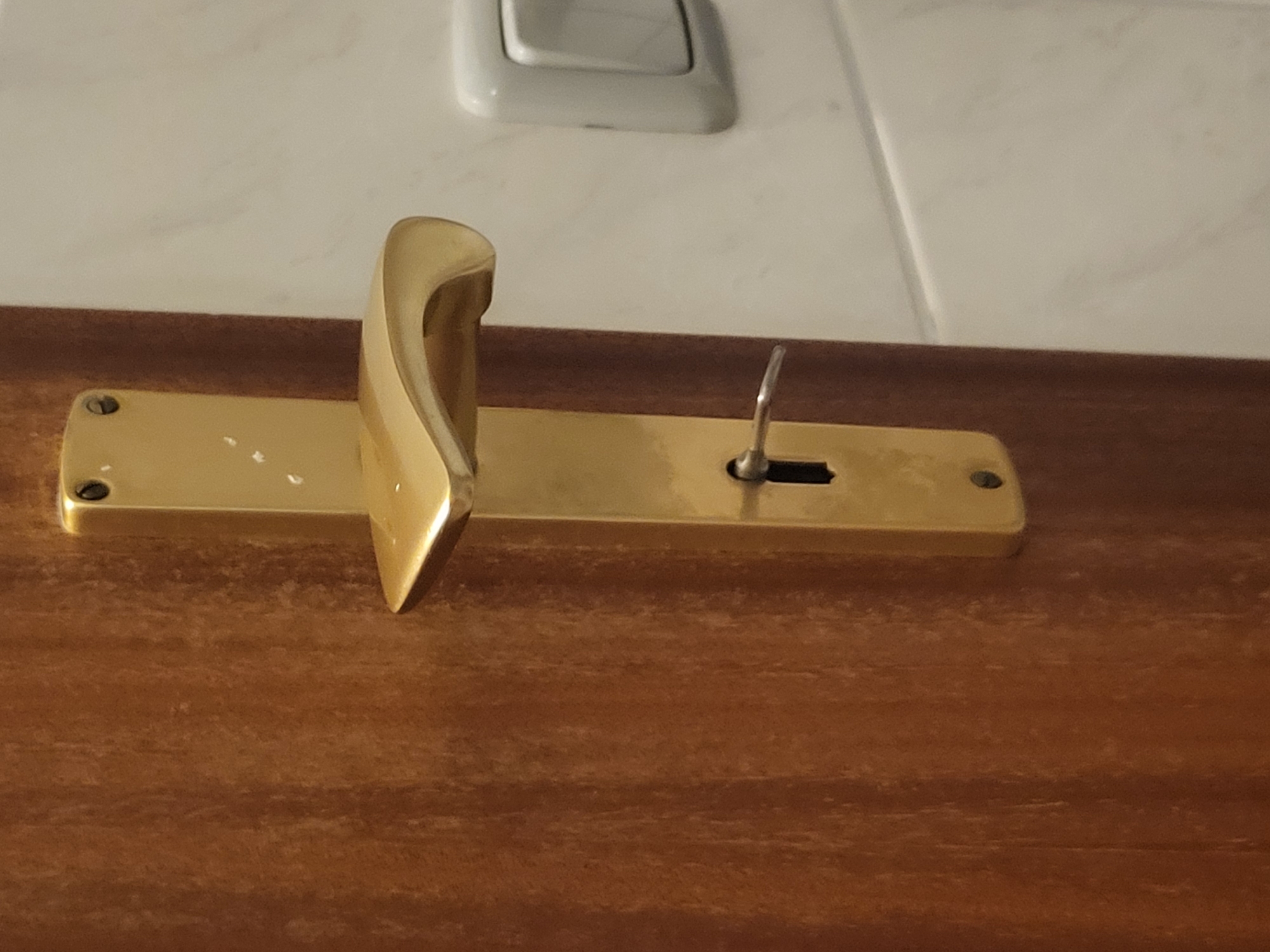 The image shows a close-up view of a golden door handle with a key inserted into the lock. The door is made of wood with a prominent grain pattern. To the right of the door handle, a white light switch is visible on a light-colored wall. The scene appears to be indoors, possibly in a residential or commercial building. There are no people present in the image. The lighting is natural and casts subtle shadows. The overall impression is one of a functional and slightly dated door mechanism. The specific location in Pinneberg, Germany, cannot be determined from the visual information provided.