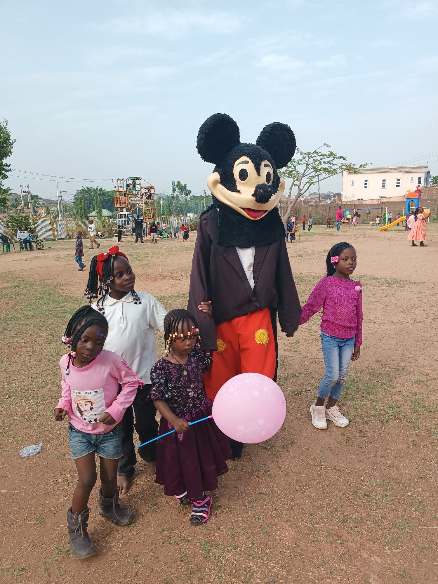 In an open, outdoor area with a dirt and sparse grass ground cover in Jos, Nigeria, a person dressed in a Mickey Mouse costume stands holding hands with four young African girls. The girls are of varying heights and dressed in colorful clothing. The girl on the left wears a pink shirt and denim shorts, the one next to her a white shirt, the girl in the middle a purple dress, and the girl on the right a pink sweater and jeans. The Mickey Mouse costume is wearing a brown suit jacket and red pants with yellow accents. The girl in the middle is holding a pink balloon on a stick.
In the background, there are people, trees, buildings, a playground, and what appears to be a ride of some sort. The sky is overcast.