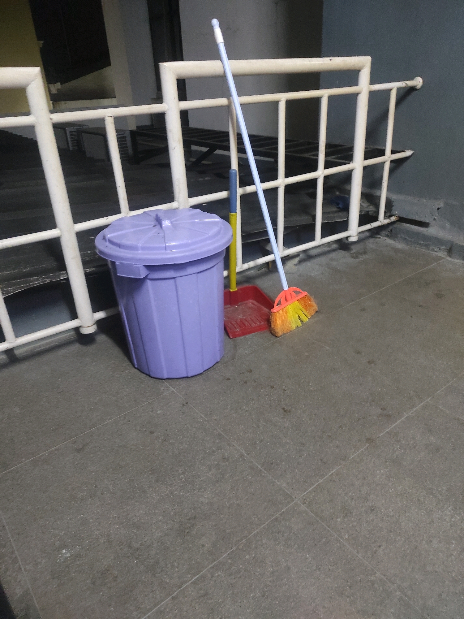 The image shows a cleaning set, consisting of a purple trash can, a red dustpan with a yellow handle, and a broom with an orange and yellow brush, placed on a tiled outdoor floor. In the background, there is a white railing and a gray wall. The scene appears to be located on an exterior walkway or balcony, possibly in a residential or commercial building. There are no people visible in the image. The setting suggests a functional area where cleaning equipment is stored. The tiles are a neutral gray color and appear to be laid in a grid pattern. The lighting suggests it might be evening or indoors with artificial light. There are some minor scuff marks and dirt specks on the floor and railing, indicating use and exposure to the elements. The trash can is cylindrical with a lid. The broom is upright, leaning against the trash can and dustpan. The dustpan is flat on the ground. The overall impression is one of an ordinary, utilitarian scene. The location is not specified within the image, and therefore it cannot be confirmed as South Jakarta, Indonesia.