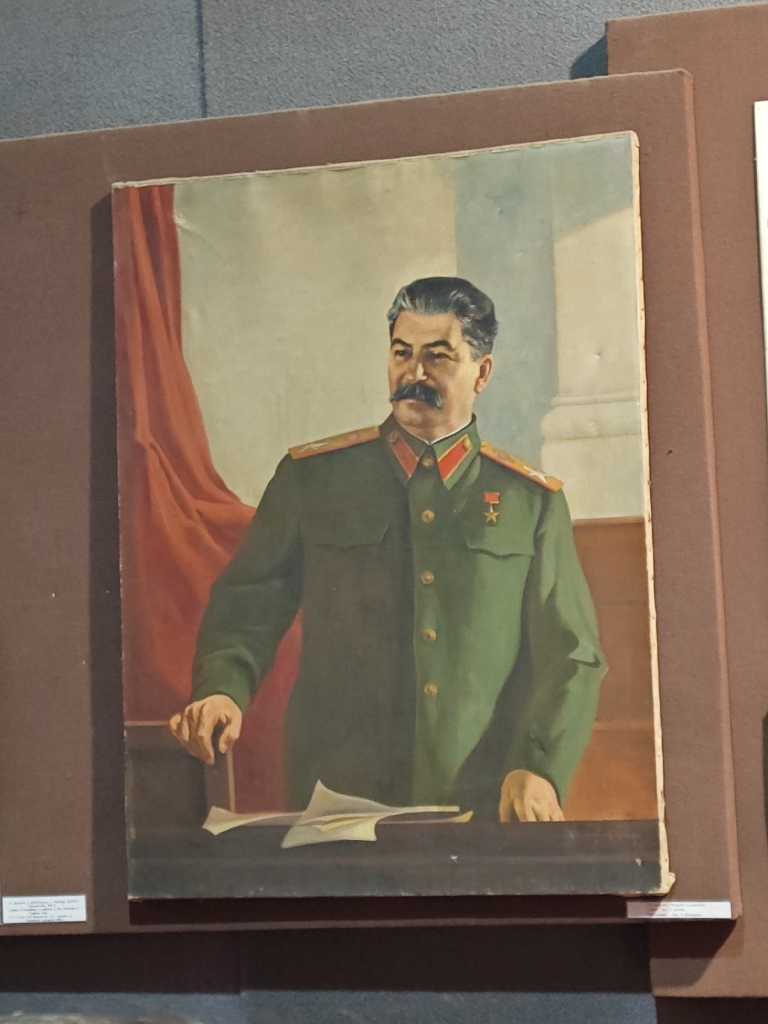 The image displays a vertical portrait painting of Joseph Stalin, presented in what appears to be a museum or exhibition setting. Stalin is depicted from the waist up, wearing a dark green military-style uniform adorned with red and gold epaulets and a red collar with gold accents. A medal featuring a gold star on a red ribbon is visible on his left breast. He has a distinctive mustache and dark hair, parted on the side, and gazes slightly to the viewer's right with a serious expression.
Stalin is shown standing behind a dark-colored desk or podium, with his left hand resting on its surface and his right hand also placed near some scattered papers. Behind him, on the left side of the painting, is a flowing red drape or curtain. To the right, pale architectural elements, possibly columns or pillars, are visible against a light blue-grey background, suggesting a formal interior setting.
The painting itself, likely oil on canvas or board, exhibits some minor surface imperfections such as slight indentations. It is mounted within a dark brown display panel, framed by a narrow light-colored border. Small, white rectangular labels are visible on the bottom left and partially on the bottom right of the brown display panel, but the text on them is too small to be legible. The overall presentation is that of a historical or propagandistic artwork on display.