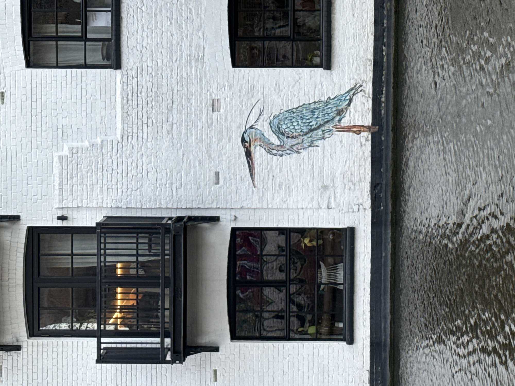 A building in London, United Kingdom is captured in this image, oriented vertically. The exterior wall is painted white, with a brick texture. There are four windows, all framed in black. One window has a fire escape structure attached to it, while another displays reflections of interior objects. Painted on the wall is a blue and gray heron. A body of water, likely a canal or river, is visible to the right of the building.