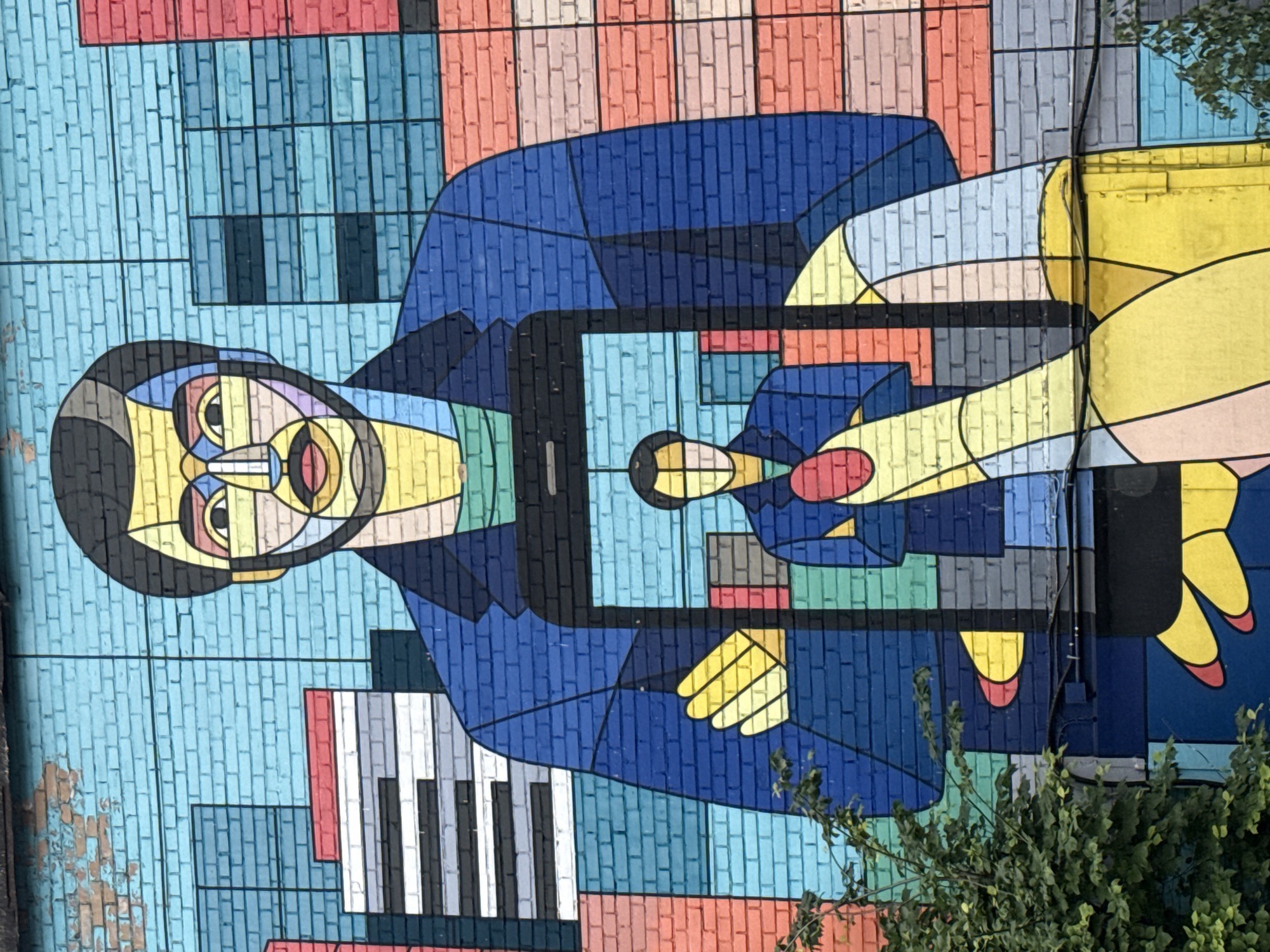 This is a close-up, eye-level shot of a vibrant mural painted on a brick wall in Moscow, Russia. The mural depicts a stylized, cubist-inspired portrait of a man, rendered in a palette of bright blues, yellows, reds, and black outlines. The man is wearing a blue jacket over a green shirt. In the center of the mural, a large smartphone screen is integrated, displaying a reflection of the same man, also stylized. The background of the mural is a geometric cityscape, featuring abstract representations of buildings in various colors, including blues, reds, and grays, mimicking the texture of the brick wall. A portion of a green leafy bush is visible in the lower left corner of the frame, partially obscuring the mural. The brickwork of the wall is visible throughout, with some signs of wear and tear, particularly in the upper left quadrant. Electrical wires are also visible running horizontally across the lower portion of the mural.