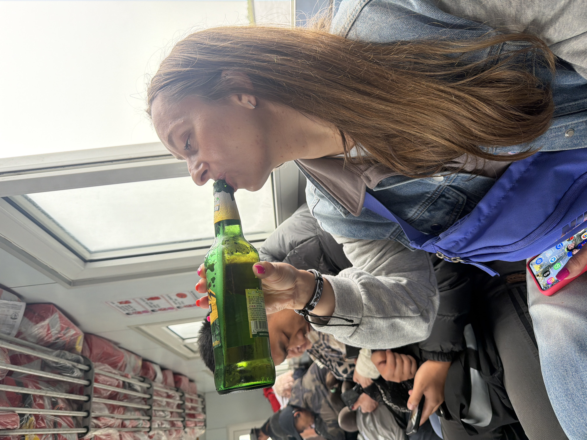 An overhead, slightly tilted perspective captures a young woman drinking from a green glass bottle within what appears to be the interior of a transport vessel or large boat, possibly a ferry, in Leninskoe, Russia. The woman, with long brown hair, wears a denim jacket over a gray hoodie and has a white wireless earbud in her left ear. Her left hand, adorned with pink nail polish, holds the bottle to her mouth. The bottle features a label with "330ml" and a partial barcode ("69299..."), along with some indiscernible Asian script.
To her right, a bright blue fanny pack or sling bag hangs, and her right hand holds a smartphone in a red case, displaying app icons and small profile pictures on its screen. She also has a patterned bracelet on her wrist. In the background, other passengers are visible but out of focus, suggesting a crowded environment.
The setting includes white-framed windows or skylights above, through which a bright, overcast sky is visible, indicating daytime. Along the left side of the cabin, rows of red-wrapped packages, possibly cargo or emergency supplies, are neatly stacked on metal racks. A partially visible red sign with white text is also present above these racks. The lighting is natural and bright from the windows. The overall scene suggests travel or transport, with the focus on the woman's casual activity of drinking.