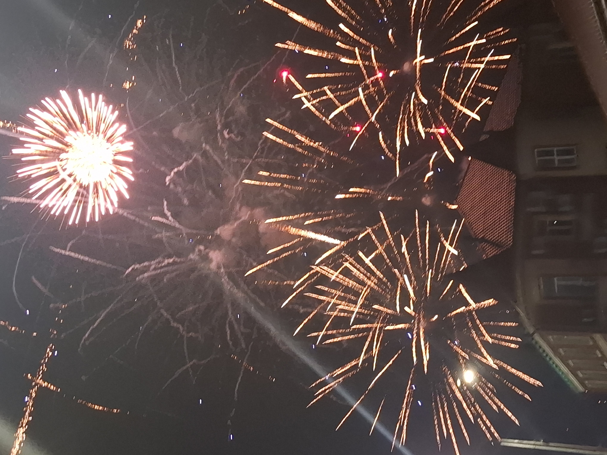 The image captures a fireworks display over the city of Svobodny, Russia. The night sky is filled with exploding fireworks, predominantly in shades of gold and red, with streaks of light extending outwards from their detonation points. Smoke trails from previous bursts are also visible. In the foreground, the roofs of several buildings are silhouetted against the dark sky, with some windows and architectural details discernible. A street lamp is visible on the left side of the image, providing a subtle illumination. The overall scene depicts a festive or celebratory event taking place in an urban setting at night.
