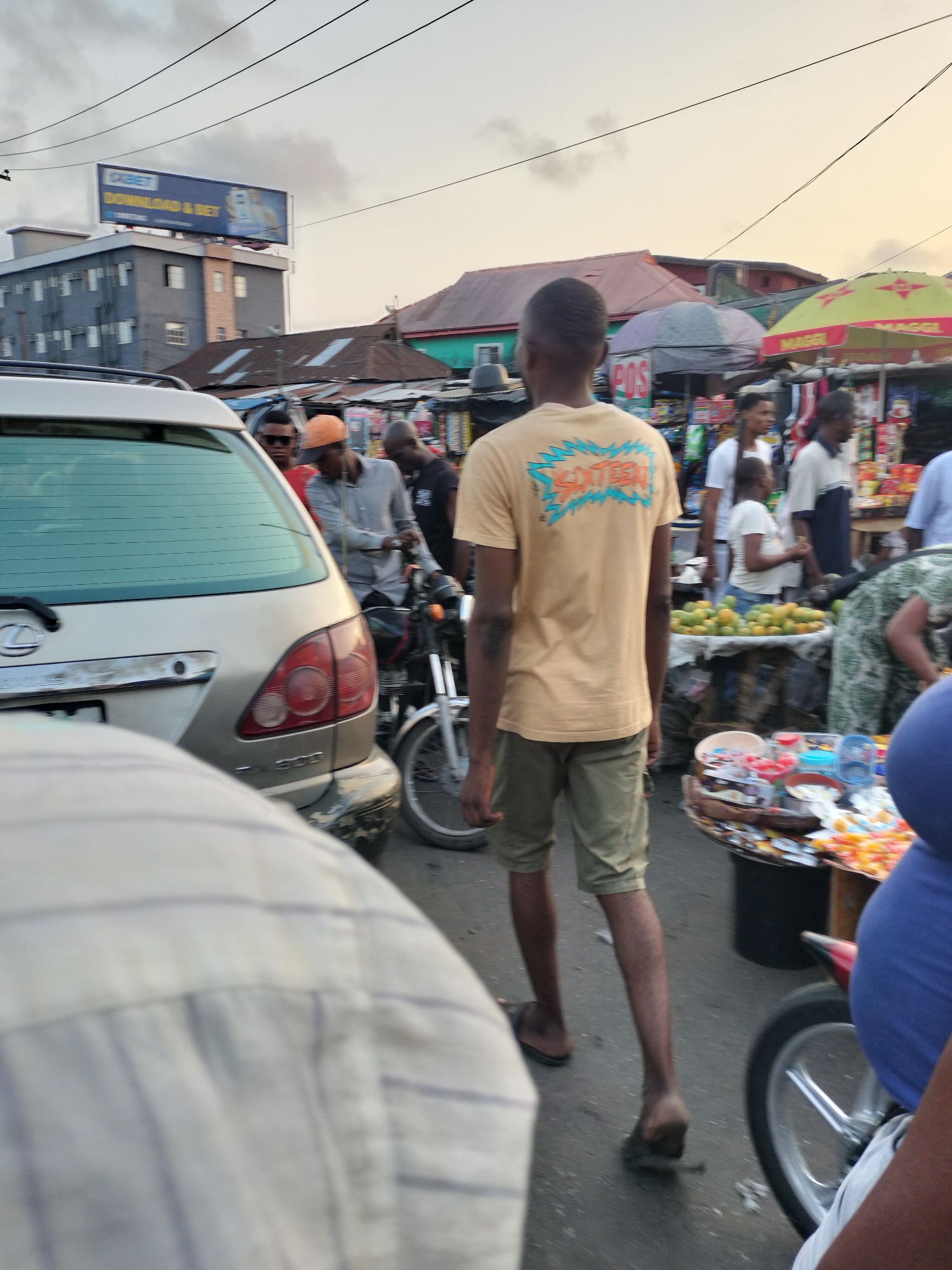 The image depicts a busy street scene in Lagos, Nigeria. In the foreground, a silver SUV with a partially visible license plate is parked. Several individuals are present. One person, with his back to the camera, wears a yellow t-shirt with the word "SXTEEN" on the back, khaki shorts, and sandals. Another person is partially visible in the bottom left corner, wearing a striped shirt. Other figures are scattered throughout the middle ground, including individuals standing near market stalls selling various items. A motorcycle is positioned near the SUV. Background elements include buildings, billboards (one advertising 1XBET), and market umbrellas. Overhead power lines are also visible.