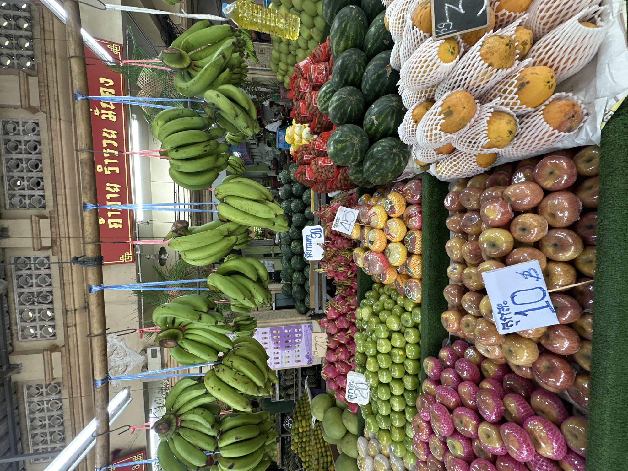 The image shows a vibrant fruit stall in Phuket City Municipality, Thailand. A variety of fresh fruits are meticulously arranged, including bunches of green bananas hanging from the ceiling, piles of green apples, red and pink apples, dragon fruit, watermelons, and papayas wrapped in protective netting. Several price tags are visible, with one indicating "10 ฿" for apples and another showing "45" for dragon fruit. The stall is set up outdoors, likely in a market, with a backdrop of a building and a red banner with Thai script. Sunlight illuminates the scene, highlighting the colorful produce. No people are clearly visible in the foreground.