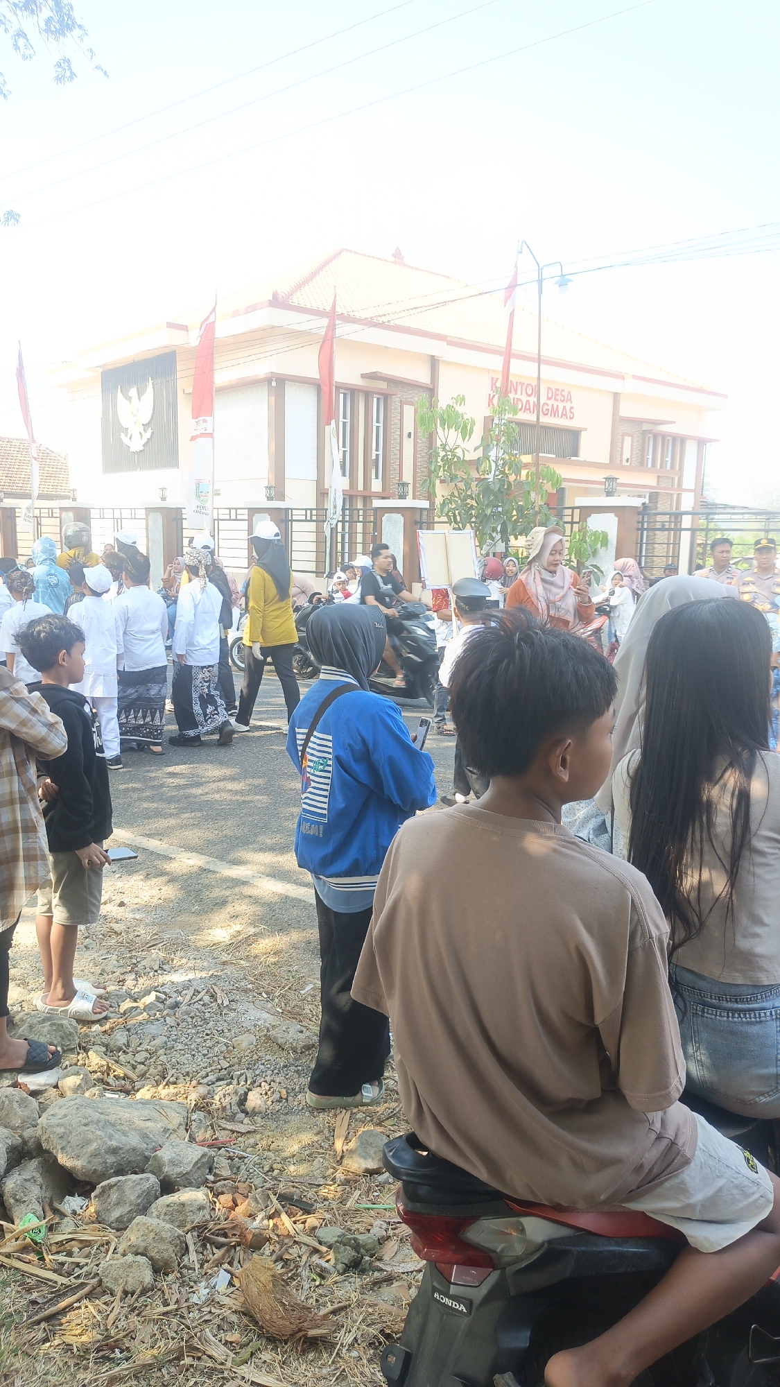The image depicts an outdoor scene in front of a building labeled "KANTOR DESA KANDANGMAS". The building appears to be a government office or community center, with a Garuda emblem prominently displayed on its facade. Several people are gathered in the foreground and middle ground, suggesting an event or activity is taking place.
In the immediate foreground, the back of a young person wearing a brown t-shirt and light-colored shorts is visible, seated on a motorcycle. To their left, another young person in a blue jacket with a graphic print is seen from the back. Further to the left, a child in a black t-shirt and shorts is also visible.
The middle ground is populated by a diverse group of individuals, including children and adults. Some are dressed in white attire, possibly uniforms, while others wear casual clothing. Several people are looking towards the building or engaged in conversation. A woman in a yellow top and headscarf is walking away from the camera. A motorcycle is also present in this area.
The environment is somewhat unkempt, with loose gravel, dry leaves, and rocks scattered on the ground. The lighting suggests it is daytime, possibly morning or late afternoon, with the sun creating a bright, slightly hazy effect. Red and white flags, likely Indonesian flags, are seen flying in front of the building. The overall impression is a community gathering in a rural or suburban setting.