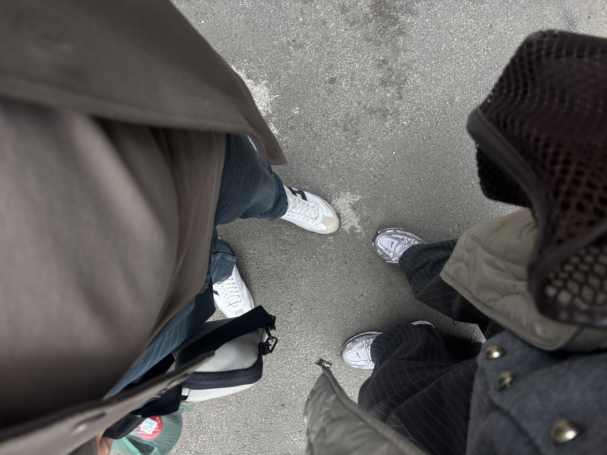 The image depicts three people standing on a grey asphalt surface. The person on the left wears a light brown coat, blue jeans, and white sneakers with a black stripe. They are holding a grey and black bag. The person on the right wears a grey jacket, a light green padded vest, and pinstripe pants. They have silver sneakers. The third person is wearing a tan trench coat and blue jeans. The background is the gray asphalt. The location of Yerevan, Armenia, is not verifiable from the image.