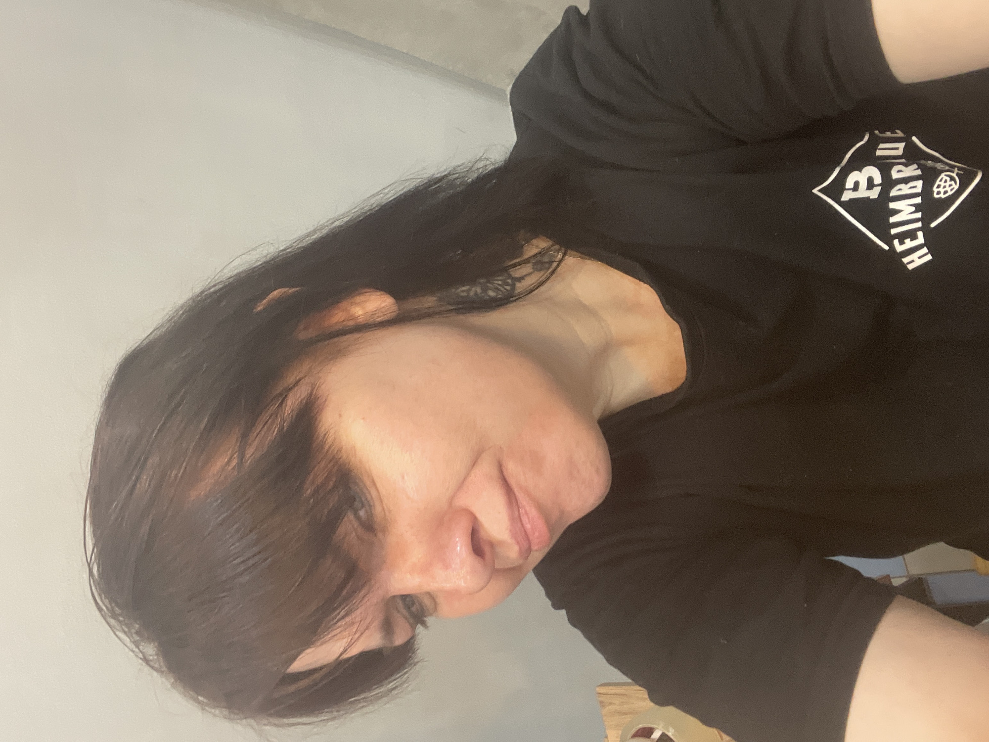 A medium close-up shot shows a woman with dark brown hair and bangs looking down and to her left, with her head tilted. She is wearing a black t-shirt with a white logo on the left side of her chest that reads "B HEIMBRUE". The background is a plain, light-colored wall. No specific location in Zhukovo, Russia can be identified.