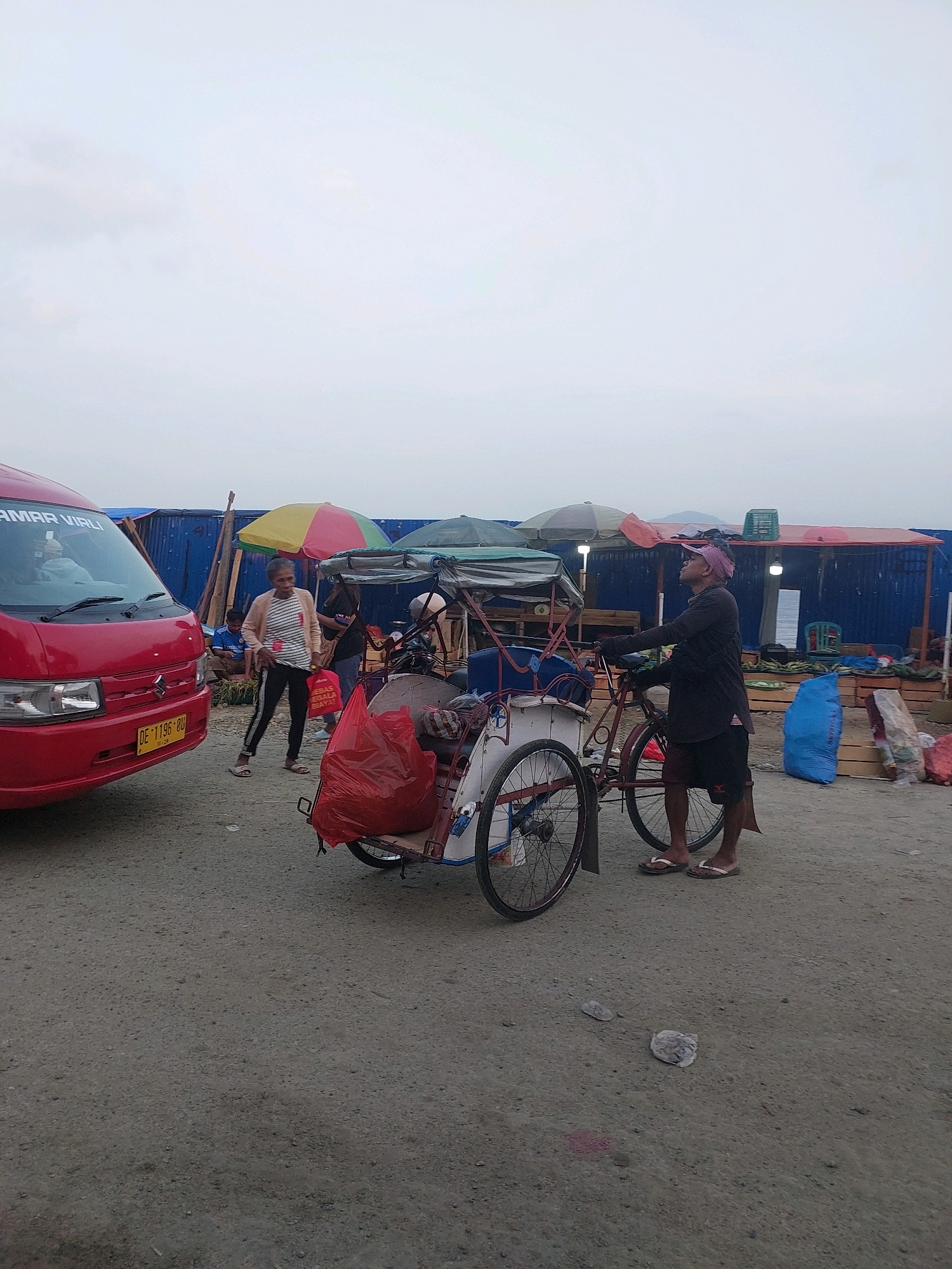 The image depicts an outdoor market scene in Ambon, Indonesia, likely during overcast weather. In the foreground, a red van is parked on a gravel surface. To the right of the van, a woman stands near a three-wheeled vehicle, possibly a pedicab, which is laden with a large red bag. Further to the right, a man wearing shorts and a dark shirt stands next to the pedicab, looking upwards. Behind them, market stalls with blue coverings and various goods are visible, including produce and large blue bags. Colorful umbrellas are set up over some of the stalls. The overall impression is one of a bustling, yet somewhat disorganized, local market.