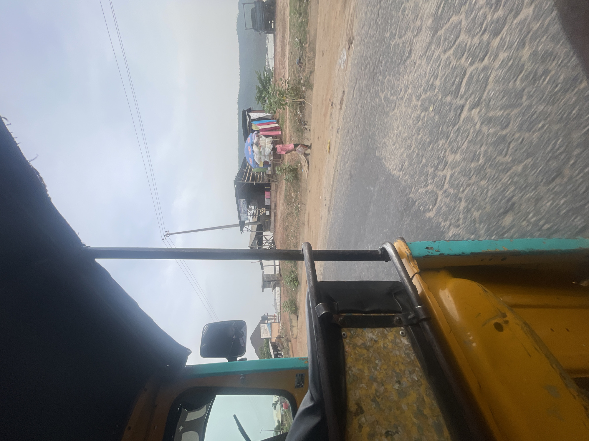 The image captures a roadside scene, viewed from inside a moving vehicle, likely a yellow and turquoise tricycle or "keke napep." In the immediate foreground, parts of the vehicle are visible, including a black fabric roof, a metal support bar, and a rearview mirror.
The scene unfolds along a straight, two-lane asphalt road that appears worn and partially covered with light-colored dust or sand, displaying a textured surface. To the right of the road is a wide, unpaved dirt shoulder with sparse green vegetation.
Along the roadside, several makeshift stalls or small shops are visible. One prominent structure features a blue umbrella, under which a person stands, possibly a vendor. Nearby, another stall displays what appears to be folded clothes or fabrics. Further down the road, more simple structures are present, and a large, dark truck is seen in the distance, either parked or in transit. Utility poles with power lines run parallel to the road, extending towards the horizon.
The background features low hills or mountains under an overcast or hazy sky, creating a muted, diffused light over the landscape. The overall impression is that of a bustling daytime environment in a semi-urban or rural area. A small, indistinct sign is visible on one of the roadside buildings, but its text is illegible.