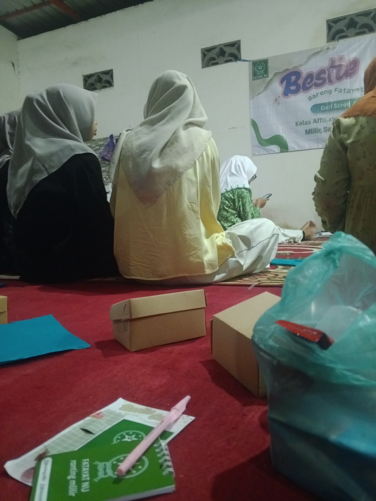 In Mlilir, Indonesia, four individuals are seated on a red carpeted floor within a dimly lit room. The person on the far left wears a grey hijab and a black garment, facing away from the camera. To their right, another individual is also facing away, wearing a light yellow hijab and garment. In the center, a person in a white hijab and green patterned top is looking at their phone. The person on the far right wears an orange hijab and a green garment. A banner with the text "Bestie Bareng Fatayat" and "Dari Scroll Kelas Affiliat Milir, Sa" is visible on the wall behind them. Various objects are scattered on the floor, including a blue paper, two cardboard boxes, a green booklet with "FATAYAT NU" written on it, and a pink pen. A teal plastic bag containing items is in the foreground.